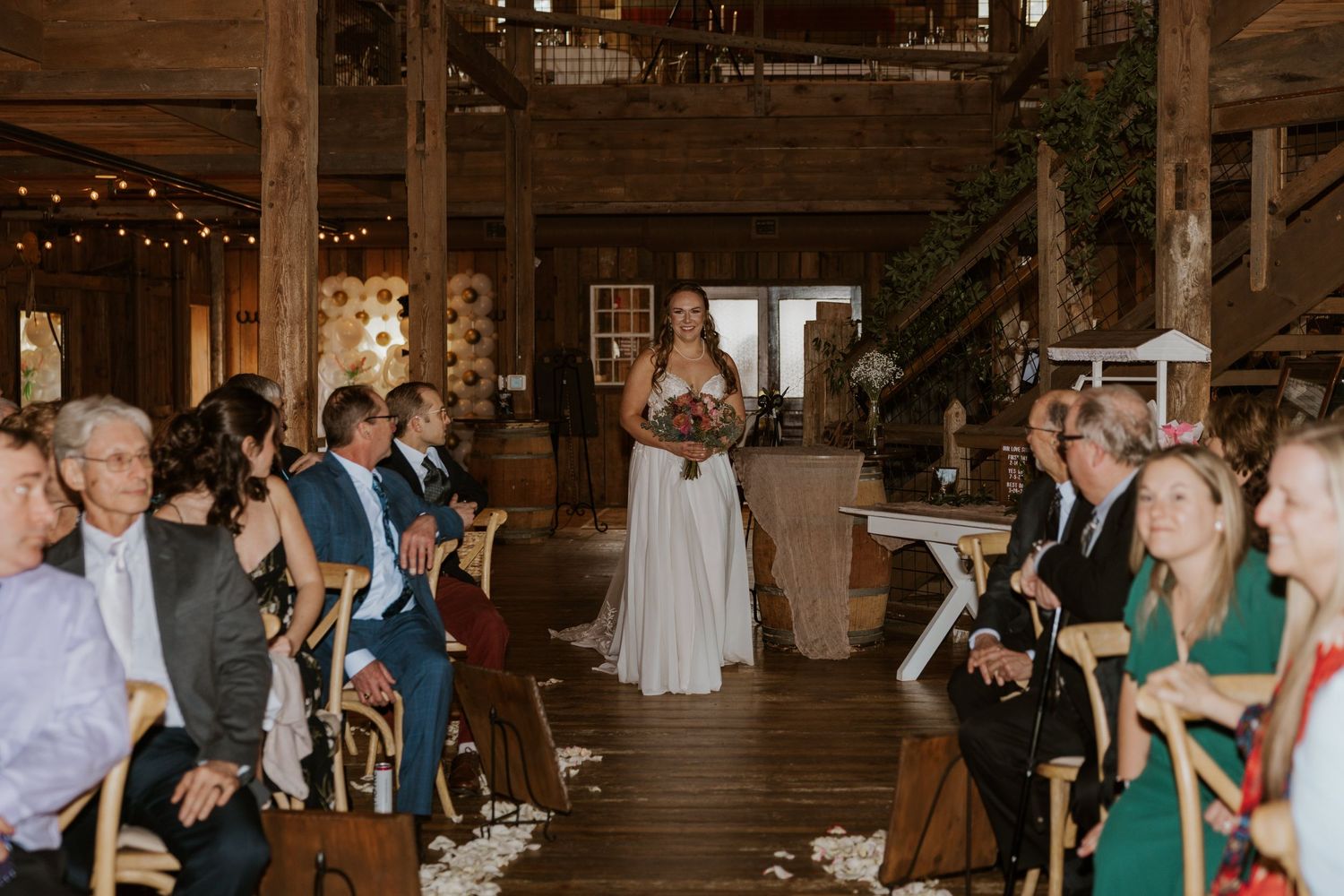 Wedding guests dance and celebrate on a rustic wooden dance floor inside a barn venue with string lights overhead.
