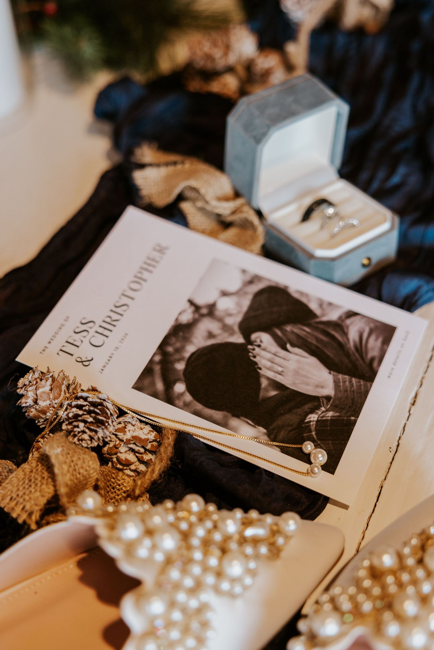 Wedding flat lay featuring Tess and Christopher program, pearl shoes, ring box, and pine cones on dark fabric.