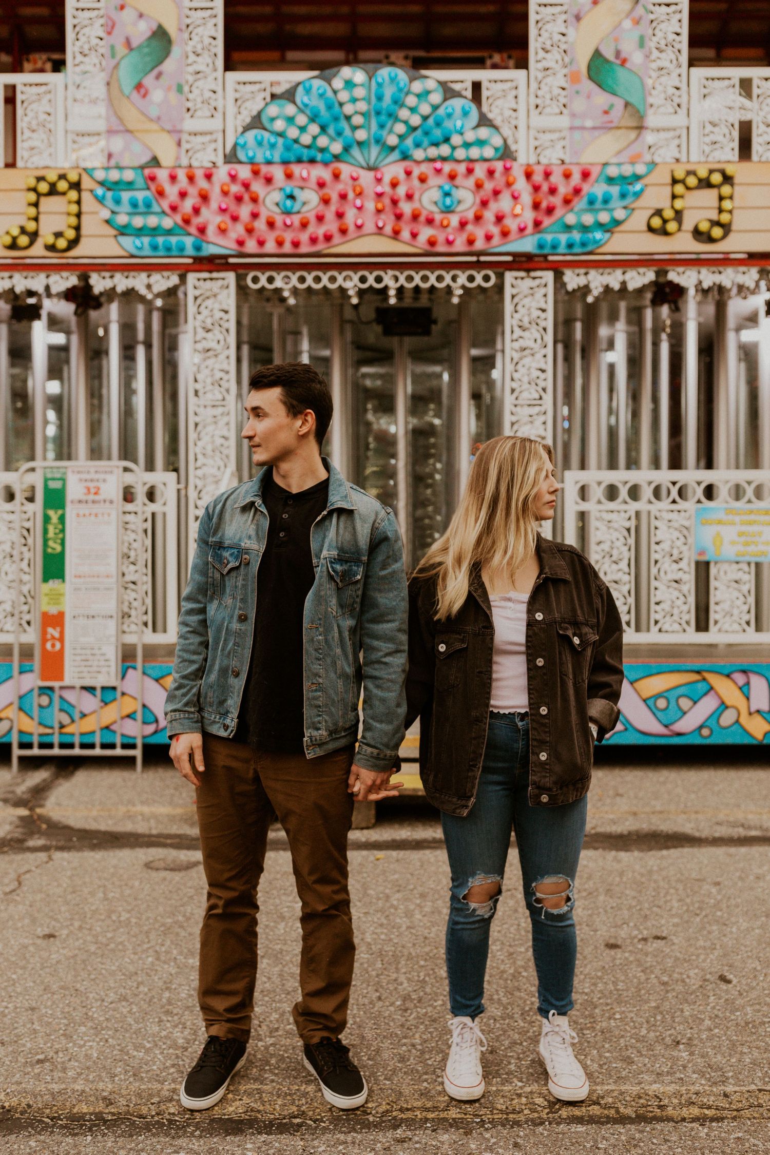 Stylish couple in denim jackets standing in front of a colorful carnival backdrop.