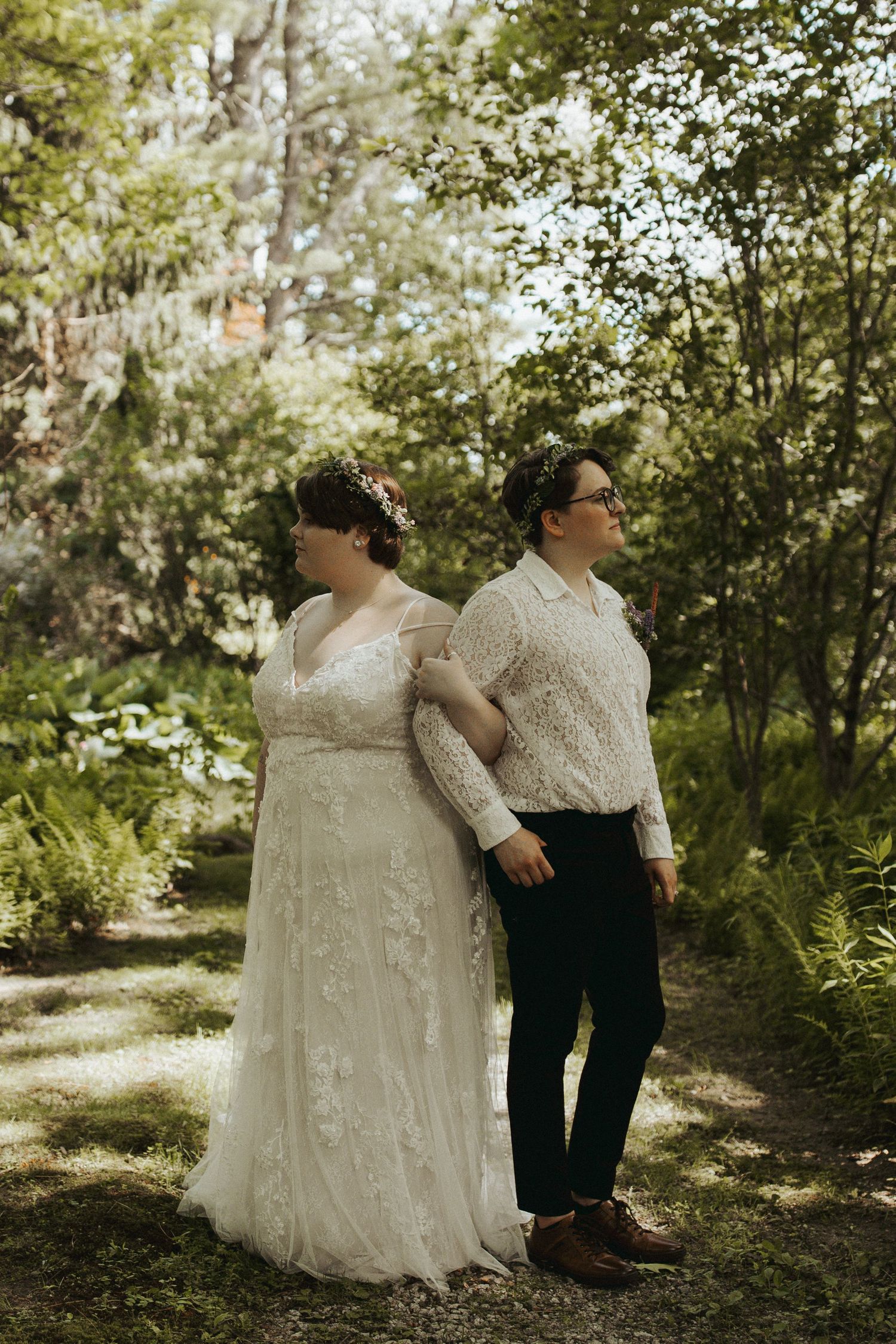 Two people in wedding attire pose together on a sunlit garden path surrounded by greenery.