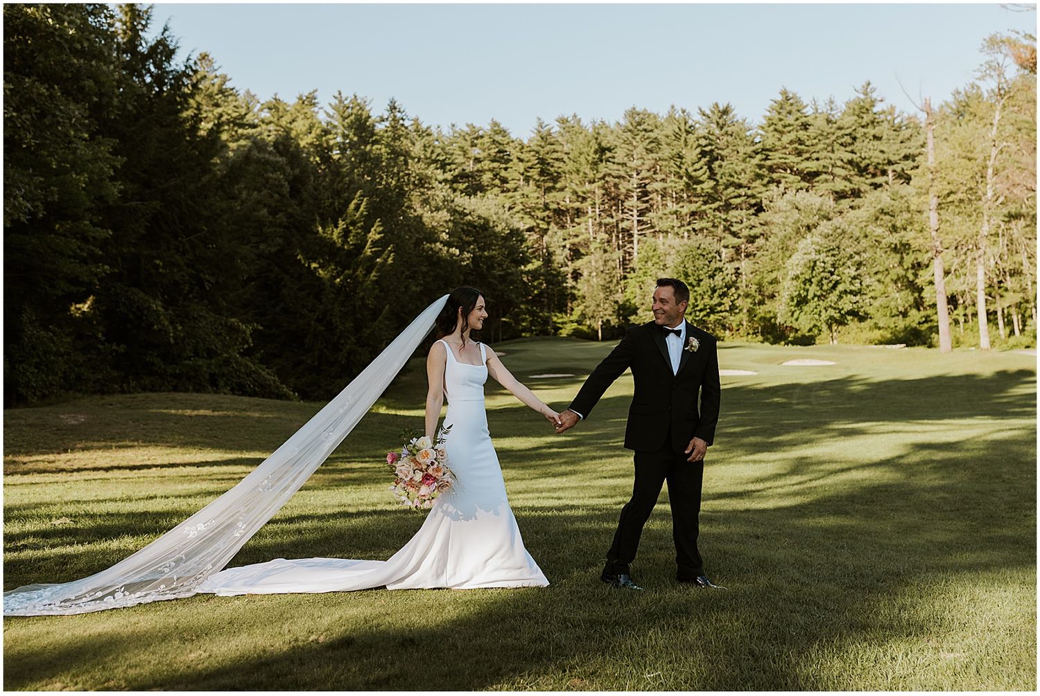 Wedding couple walking through a scenic outdoor venue with pine trees and green lawn.