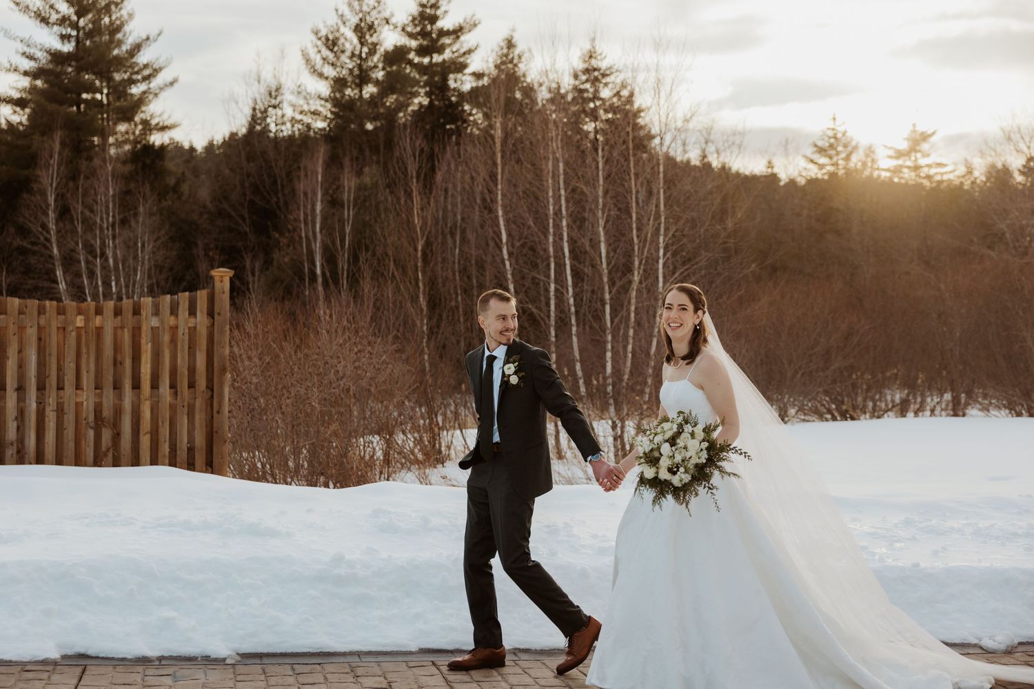Bride and groom walk hand in hand along snowy path at sunset with wooden fence and evergreens.