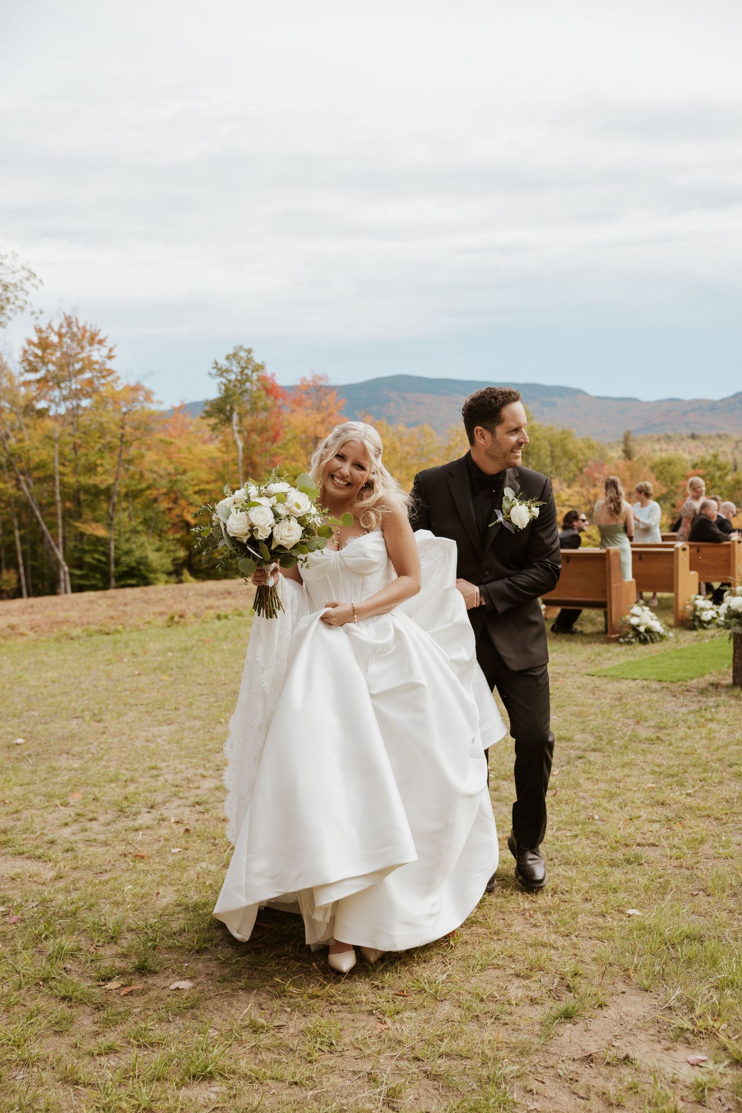 Bride in white ball gown and groom in black suit walk down outdoor aisle with fall foliage and mountains behind them.