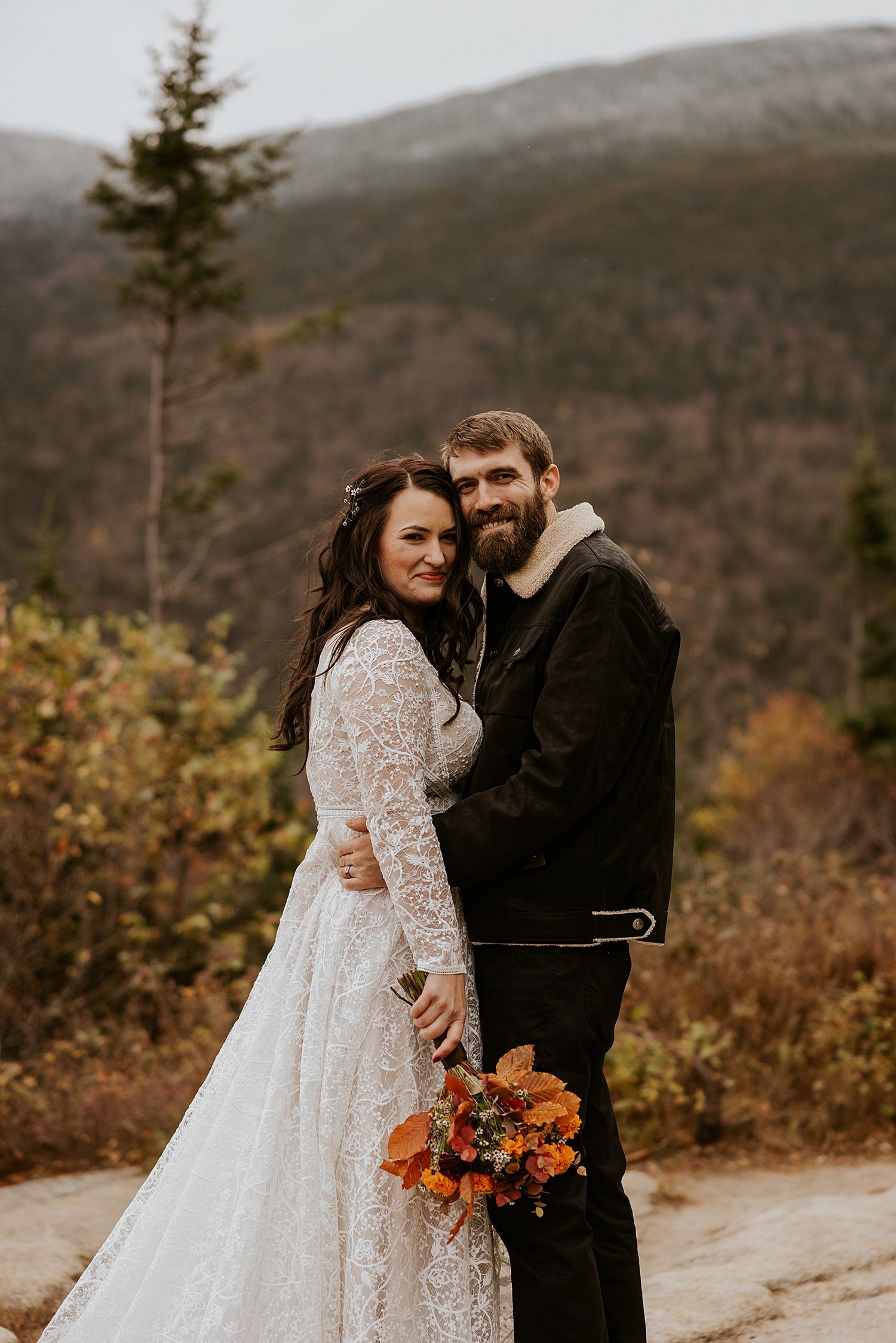 Bride in lace gown and groom in black jacket embrace outdoors with fall foliage and mountain backdrop.
