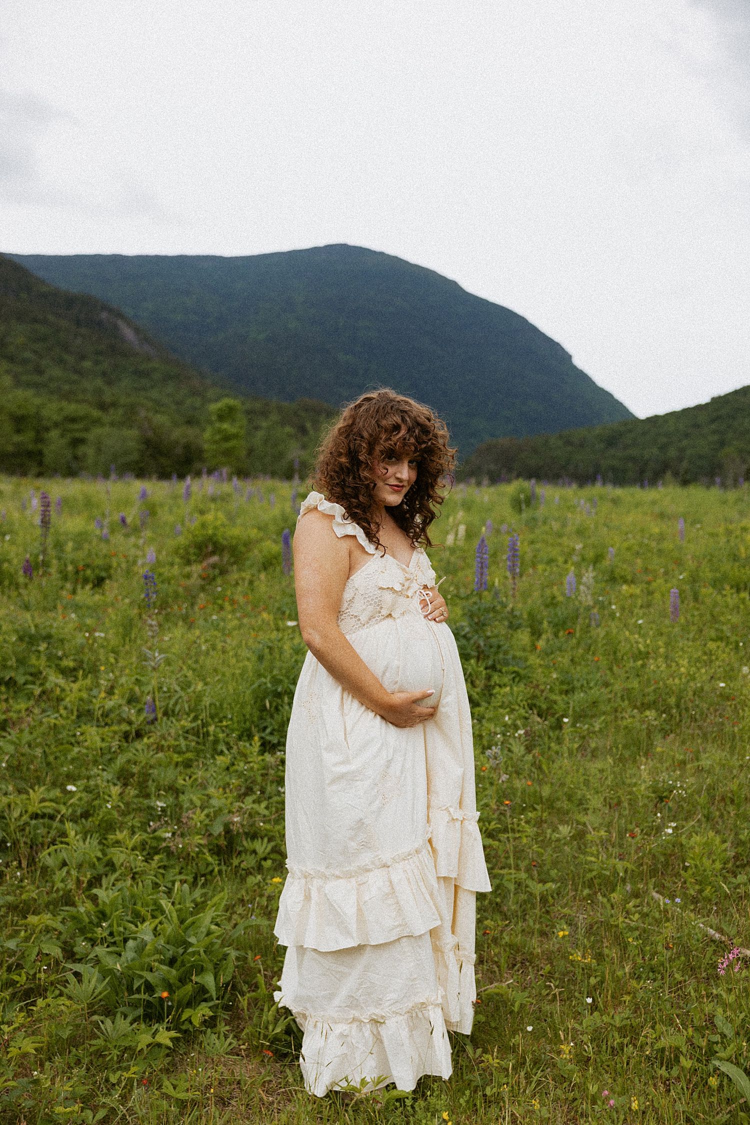 Pregnant woman in white ruffled dress standing in wildflower field in the White Mountain in New Hampshire taking motherhood photos