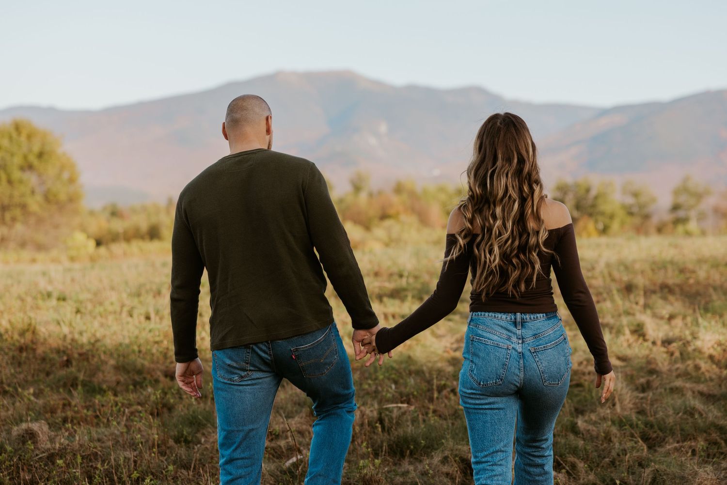 Couple holding hands walking toward mountain landscape in autumn field.
