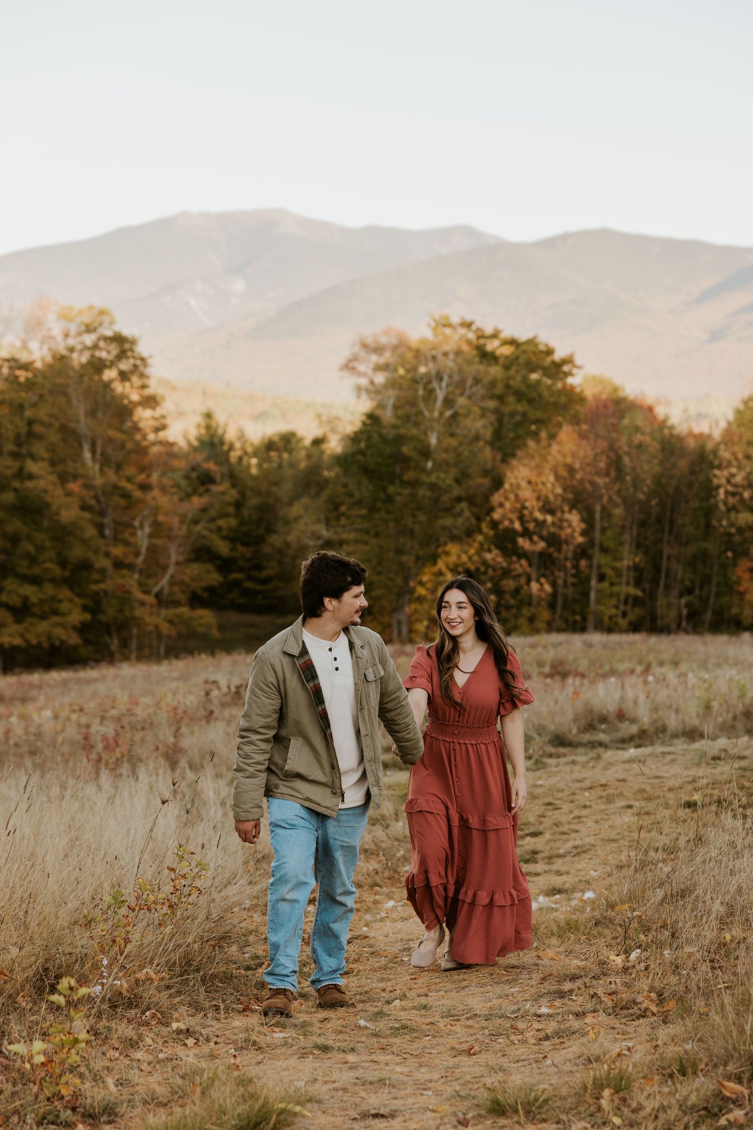 Happy couple walking hand-in-hand through a fall field with stunning mountain and autumn foliage backdrop.
