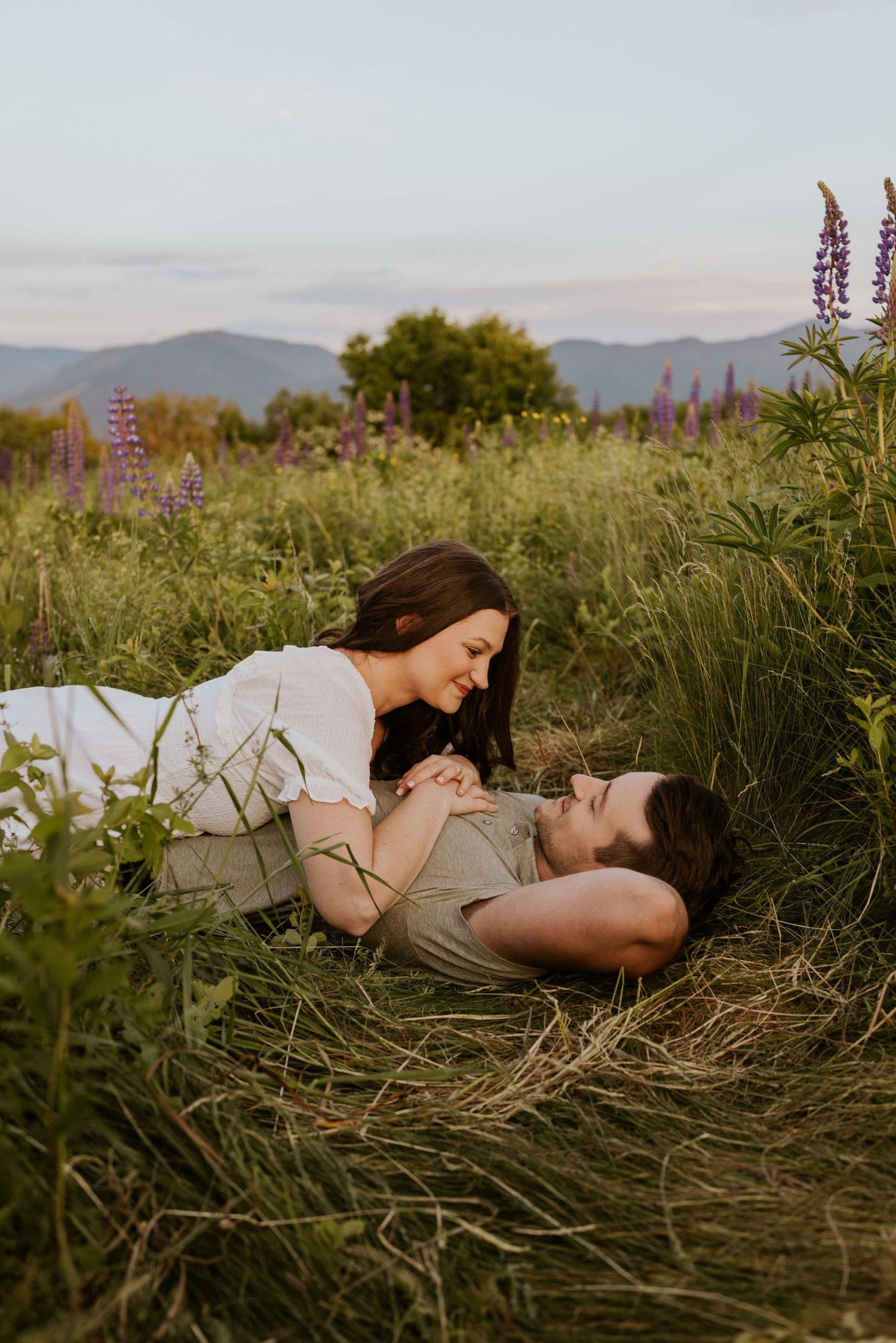 Romantic couple lying in a wildflower meadow holding hands with mountain views at golden hour.