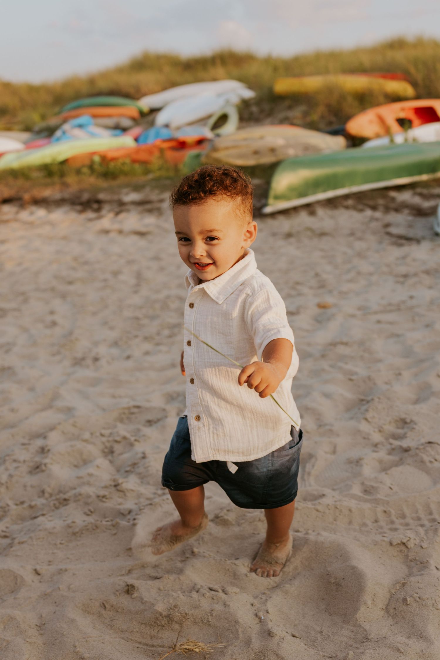 Happy toddler boy in white shirt and shorts smiling on beach with colorful kayaks behind him.
