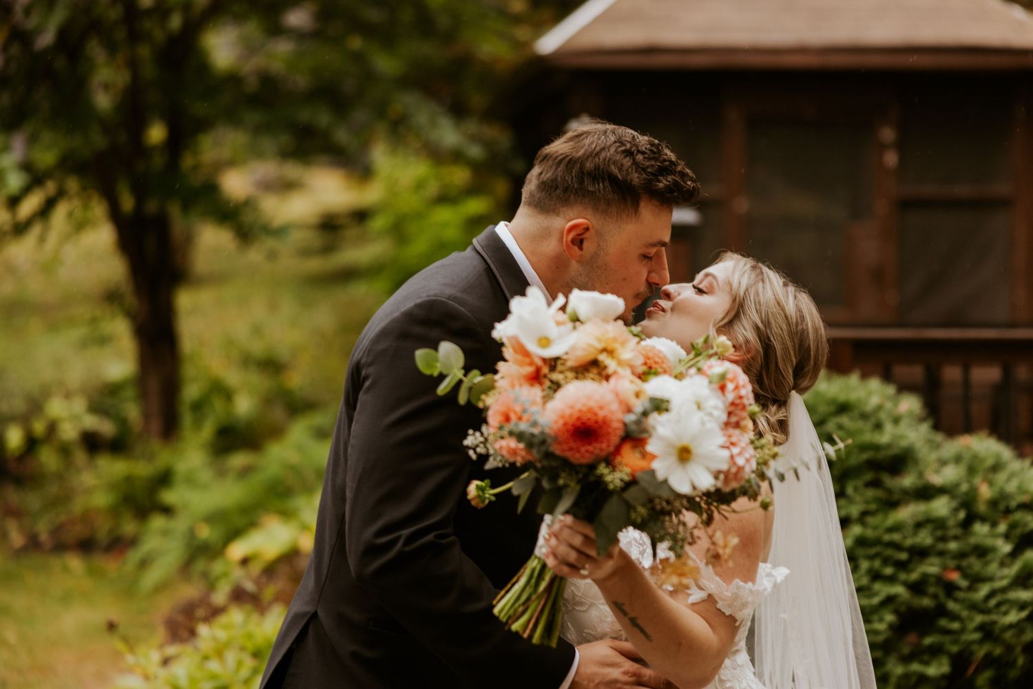 Bride and groom share a tender kiss during first look while bride holds vibrant peach and white bouquet.
