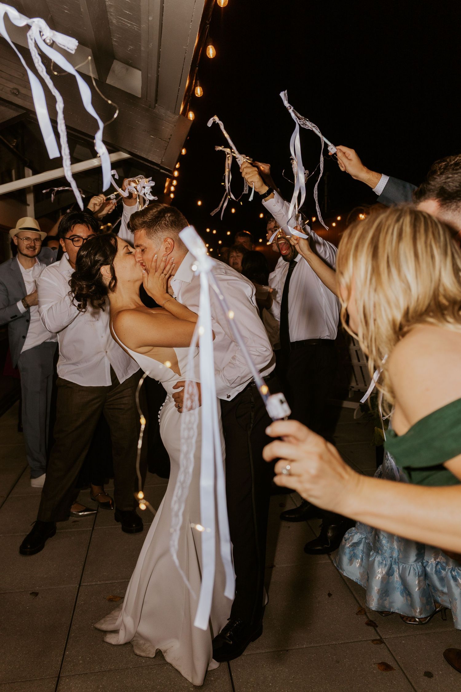 Newlyweds share a romantic kiss during ribbon wand sendoff surrounded by celebrating wedding guests.