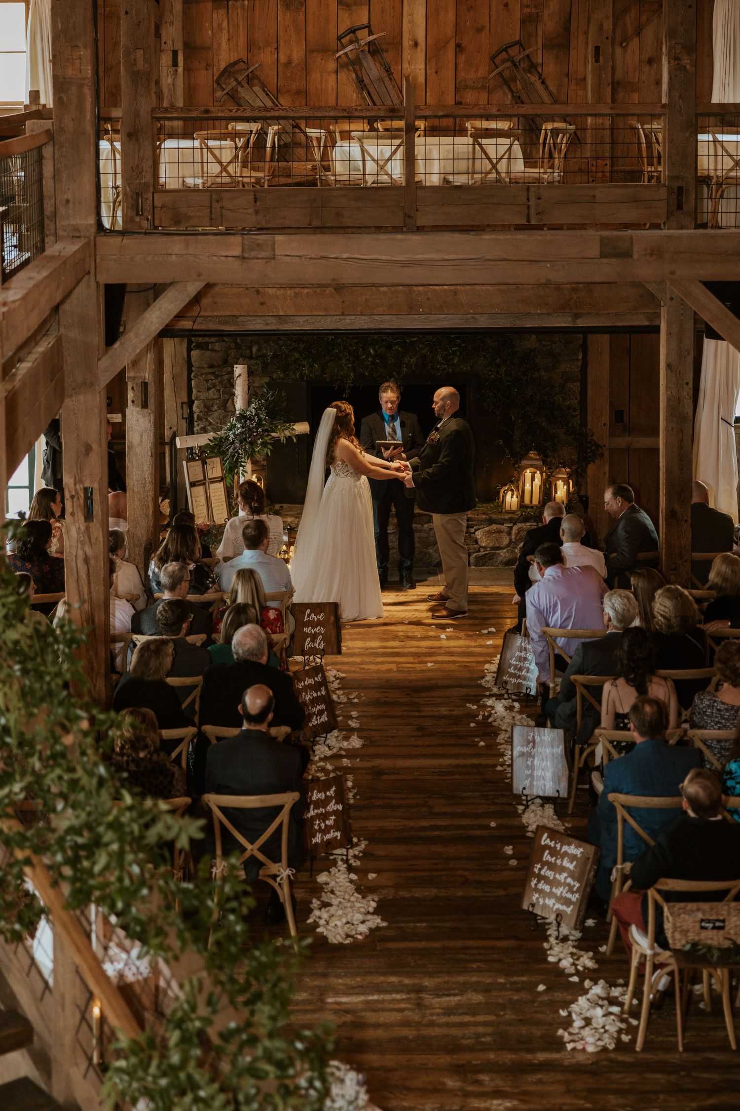 Wedding guests seated in wooden chairs inside a rustic barn venue with decorative lighting.