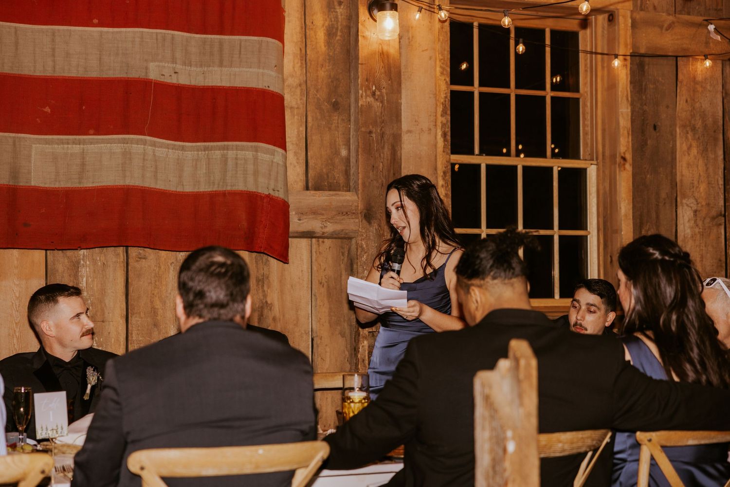 Woman giving a toast speech at a rustic barn wedding reception with string lights and American flag backdrop.