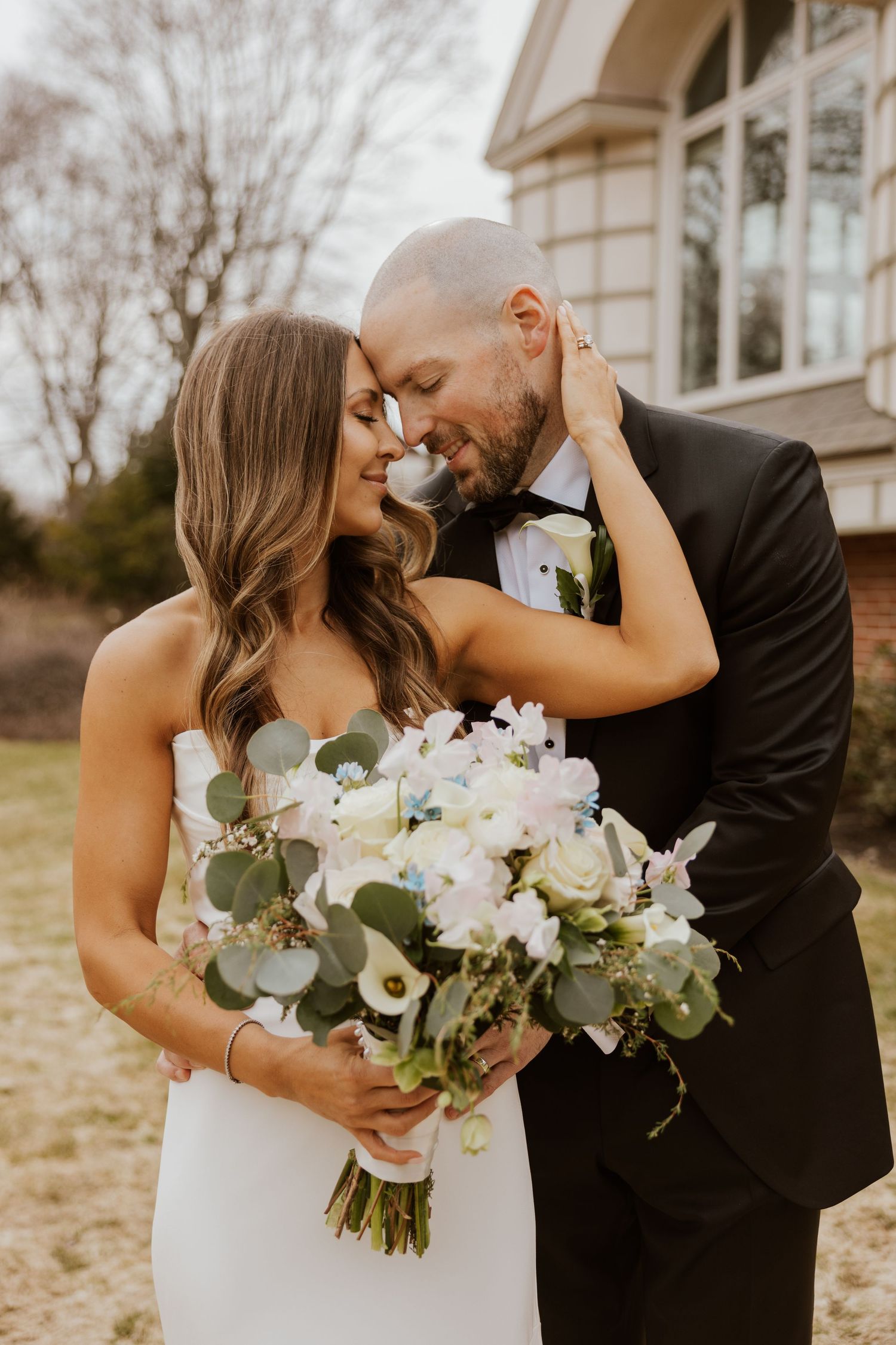 Romantic wedding couple embraces while holding white floral bouquet in outdoor setting.