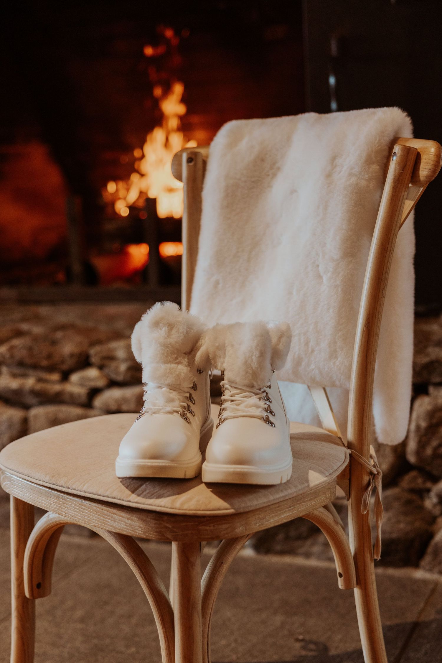 White fur-trimmed winter boots on wooden chair by cozy fireplace with fluffy white blanket.