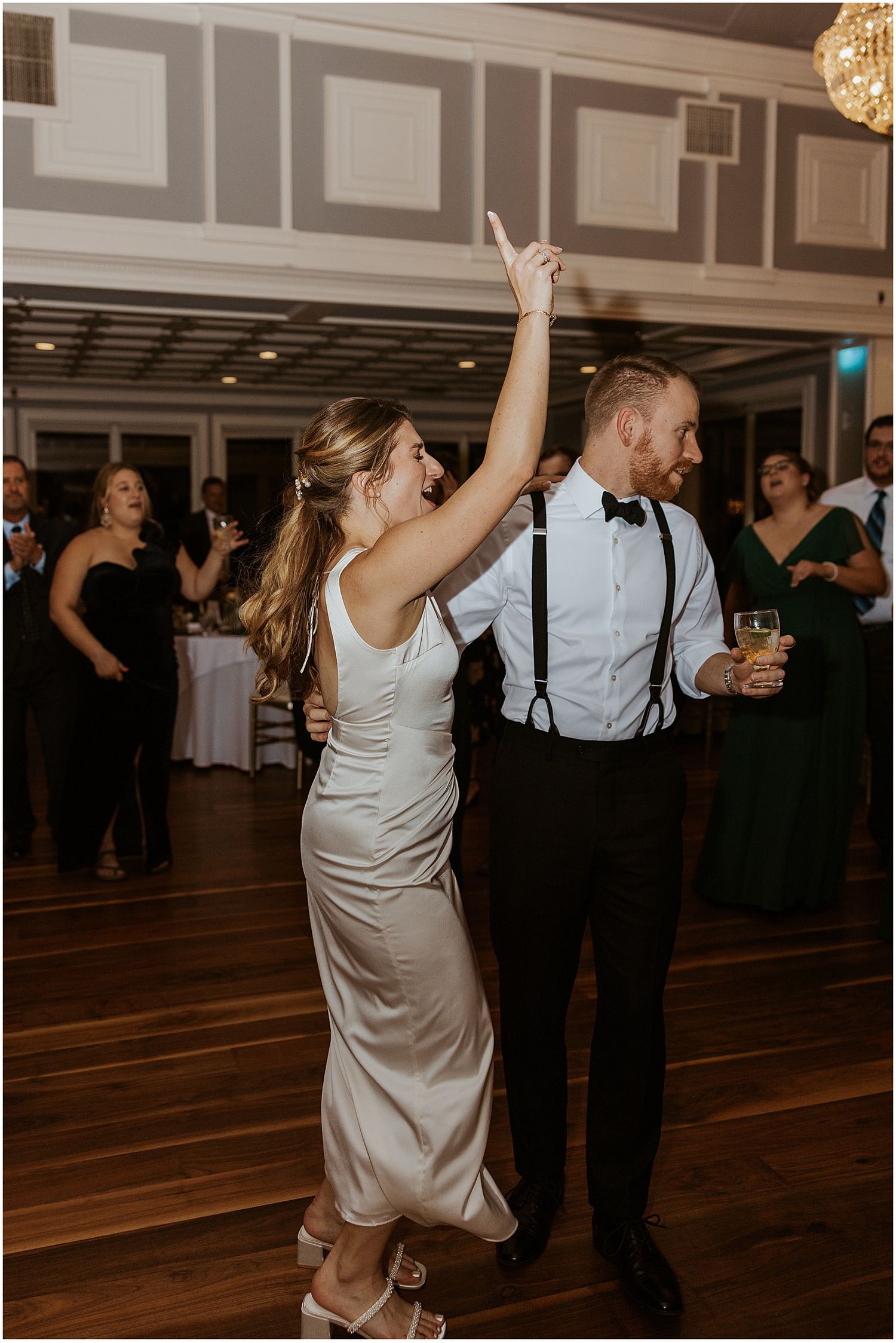 Wedding guests dancing and celebrating on a wooden dance floor at an indoor reception.