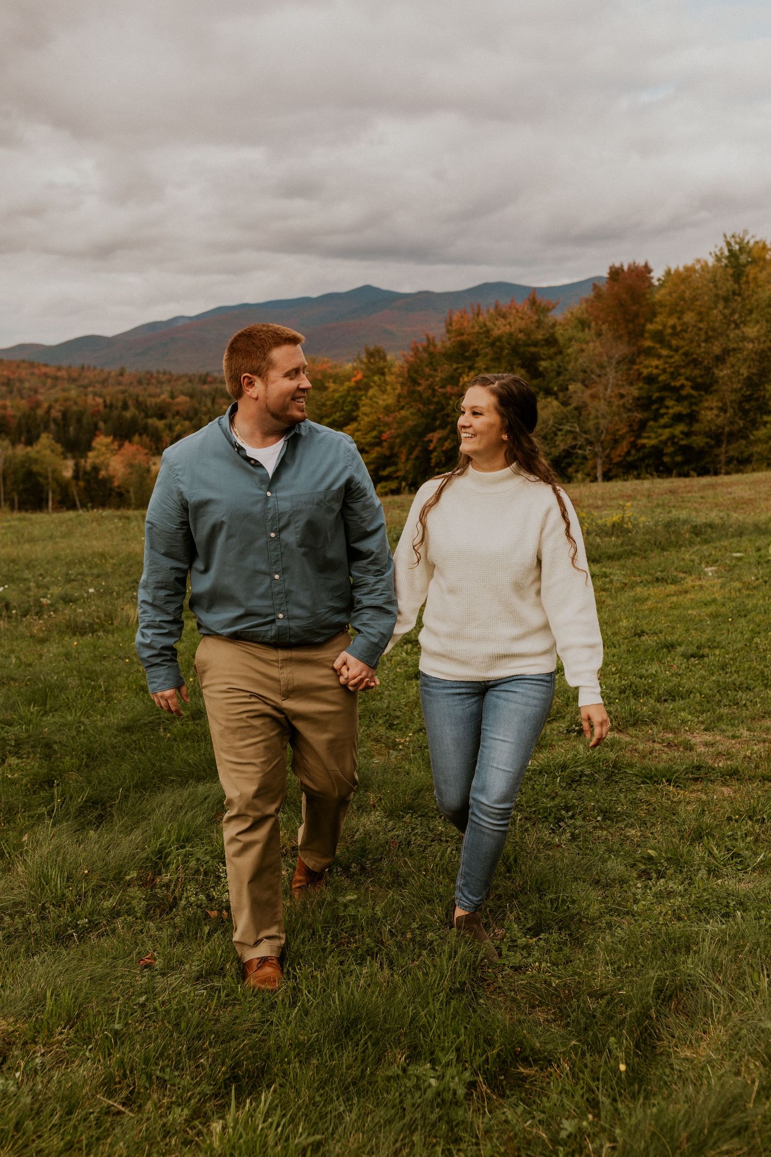 Paige and Michael walking and holding hands in a giant field with a beautiful mountain view during the NH fall foliage.
