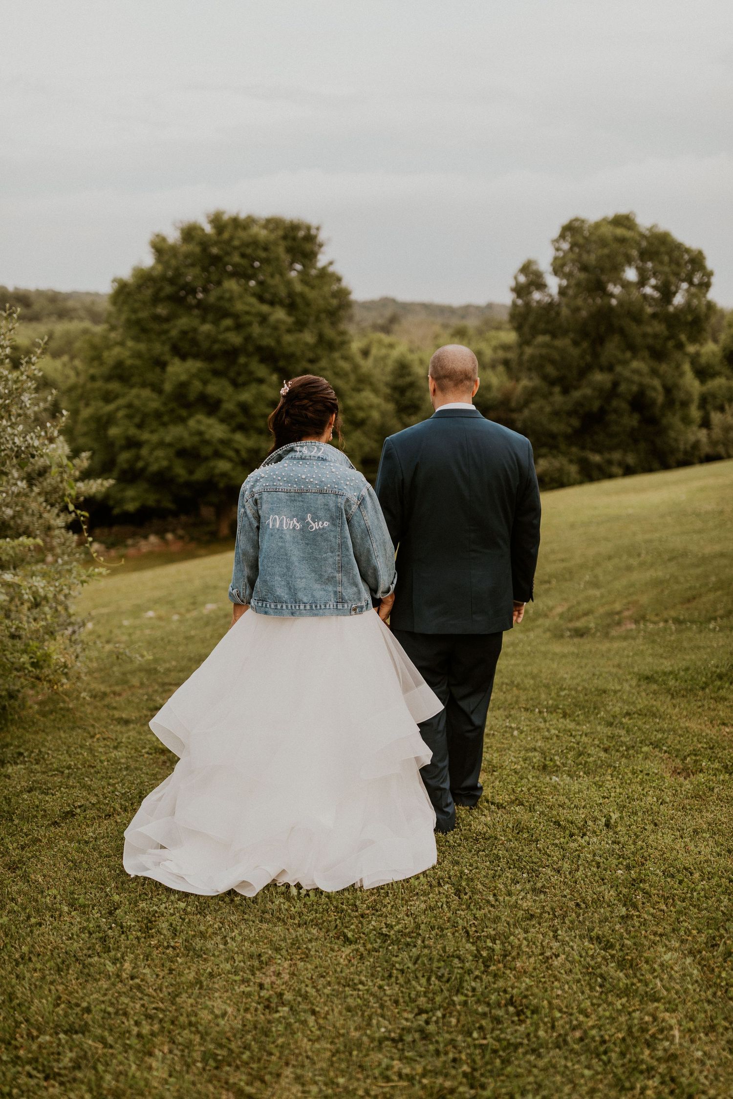Newlyweds walk together on a grassy hill with the bride wearing a denim jacket over her wedding dress.