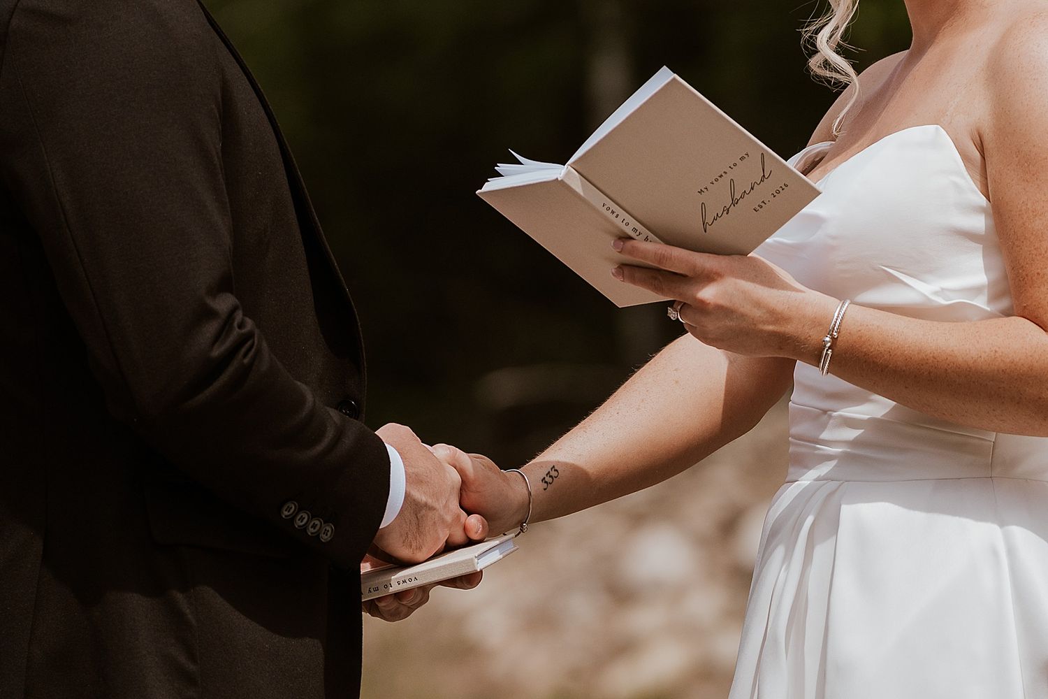 Bride and groom holding hands during outdoor wedding ceremony, bride reading vows from a small book.