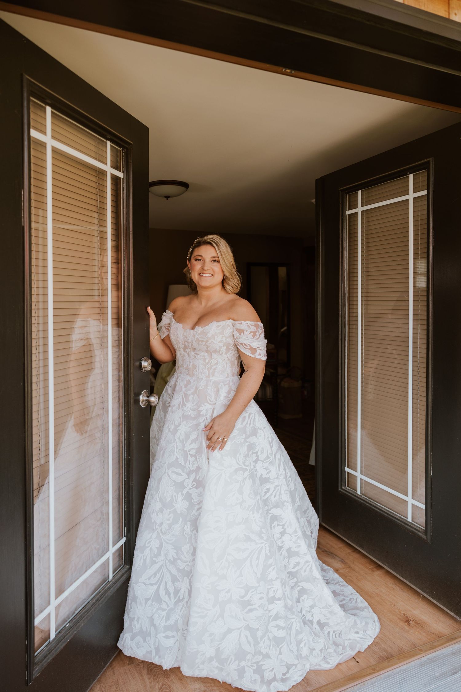 Smiling bride in an off-shoulder lace wedding gown standing in a doorway with warm interior lighting.