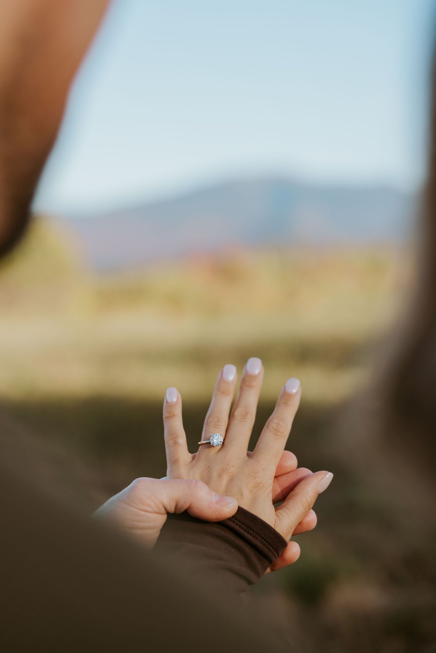 Close-up of a woman's hand displaying a diamond engagement ring held by her partner outdoors.