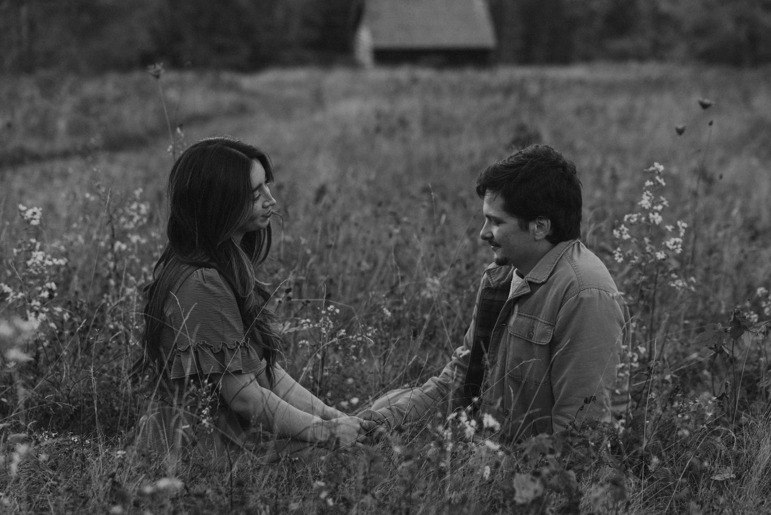 Black and white photo of couple sitting face-to-face holding hands in a wildflower meadow at dusk.