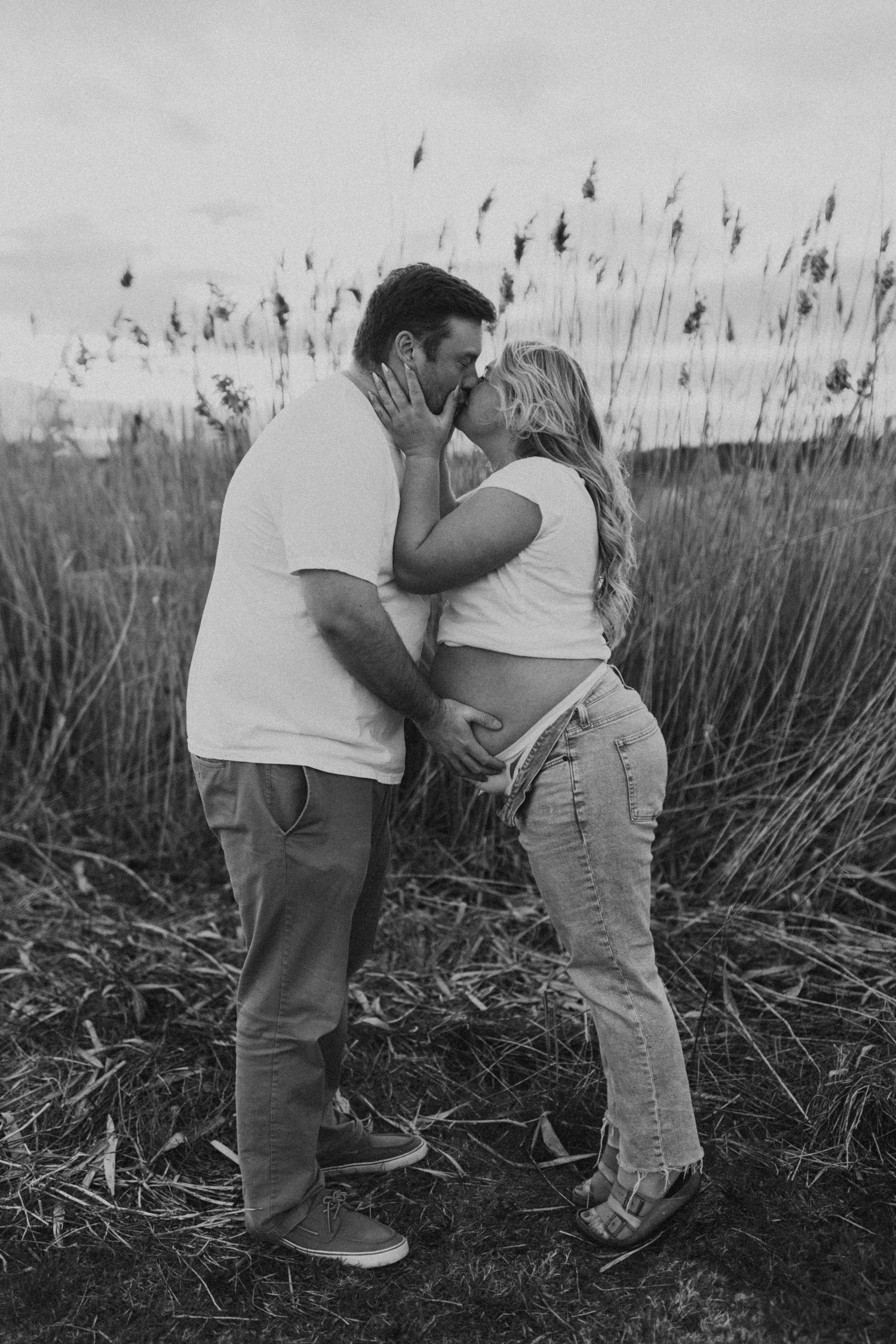 Black and white photo of expecting couple kissing in tall grass field at dusk.