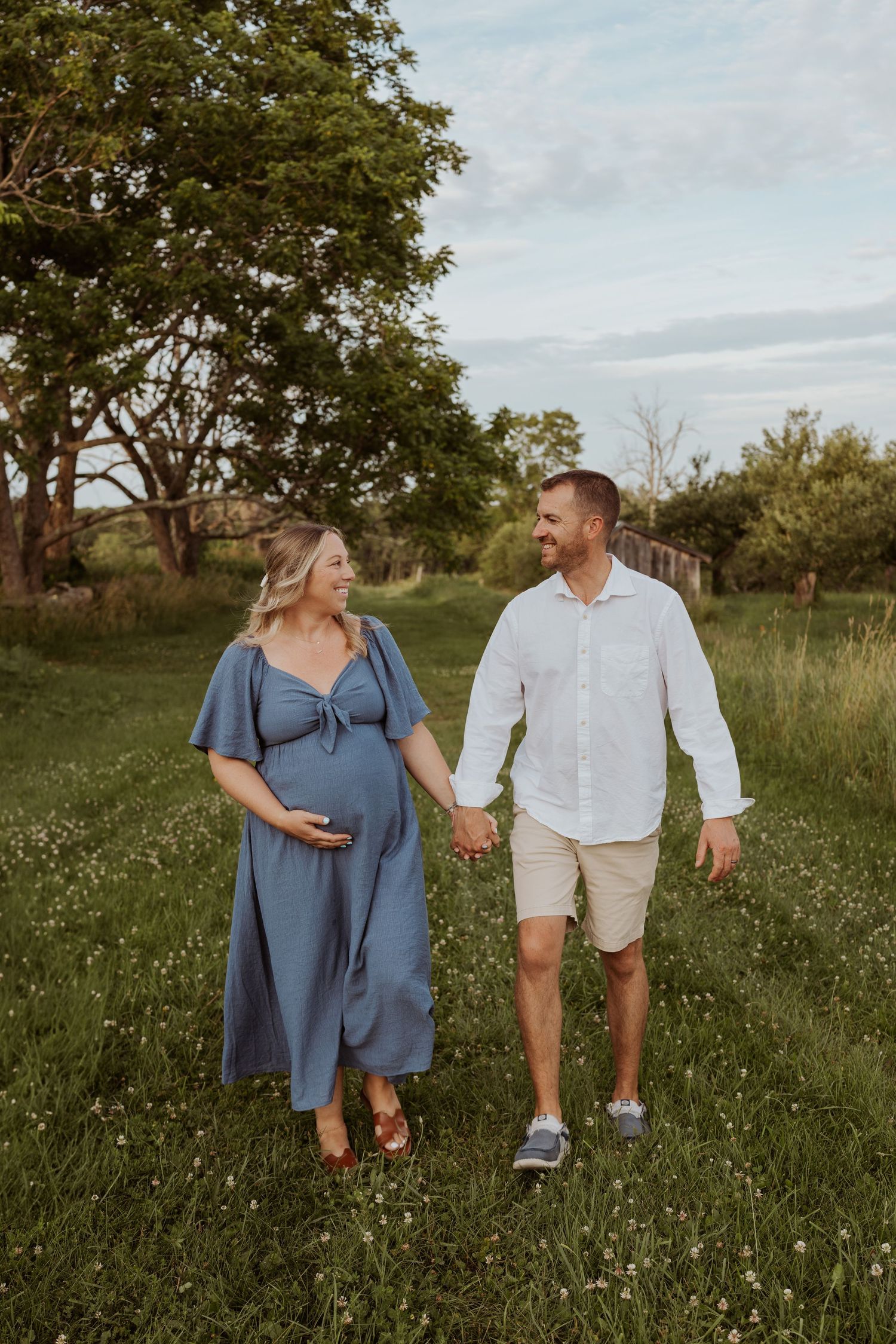 Smiling pregnant woman in blue dress holds belly while walking hand in hand with partner in field.
