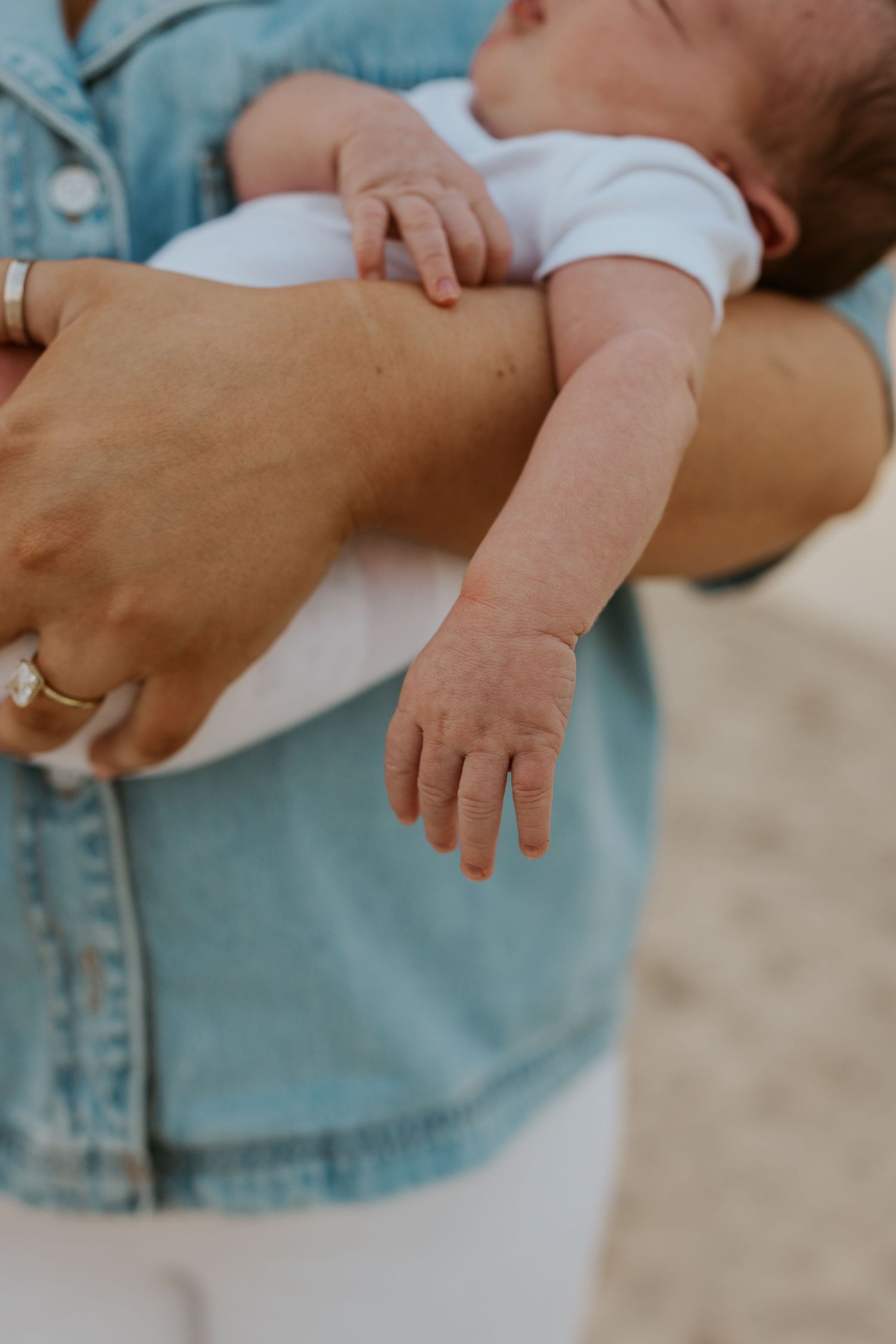 Close-up of newborn baby being cradled in mothers arms wearing denim jacket on beach.
