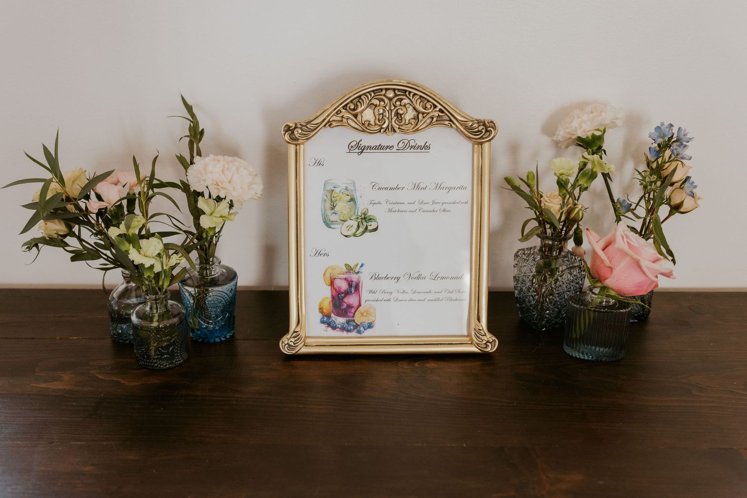 Elegant gold framed signature drinks menu display surrounded by colorful floral arrangements in blue glass vases on wooden table.