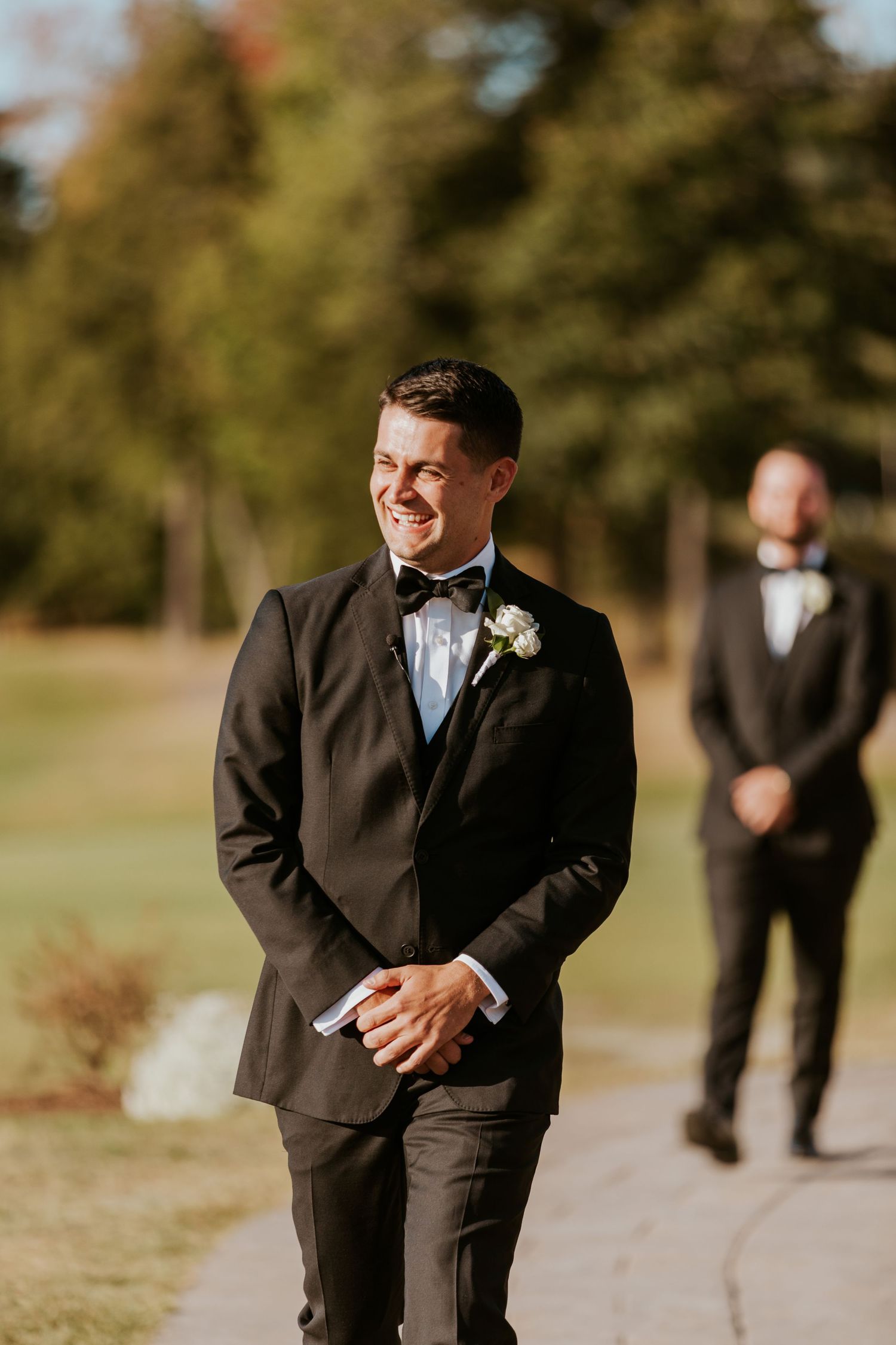 Groom in black tuxedo smiling joyfully while waiting at the altar during outdoor wedding ceremony.