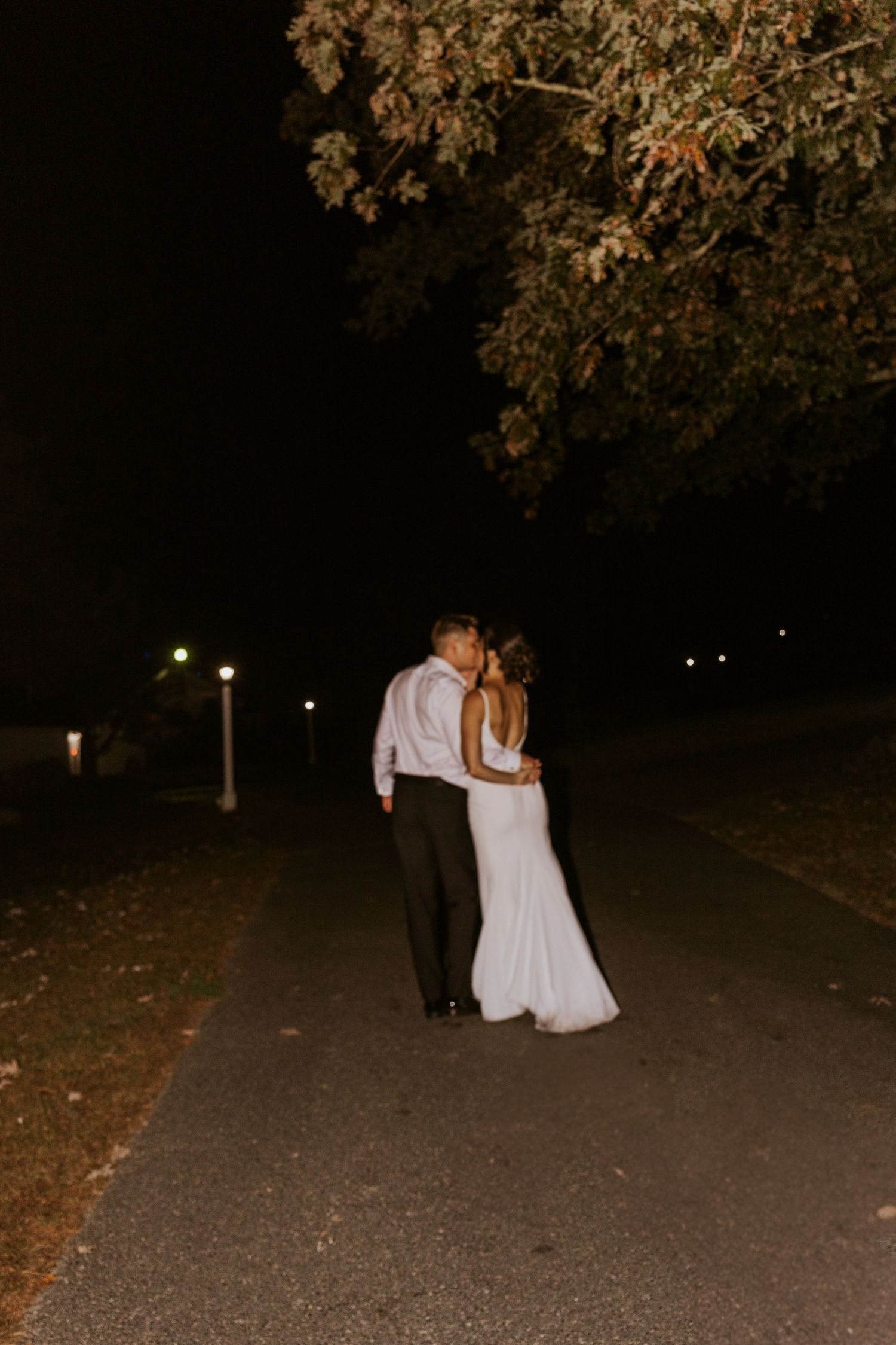 Bride and groom share a tender embrace on a dimly lit outdoor pathway surrounded by autumn trees.