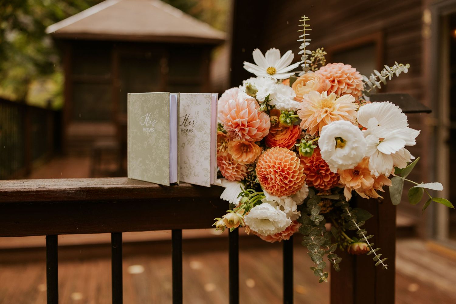 Vibrant bridal bouquet of peach dahlias and white cosmos rests beside vow books on rustic wooden railing.