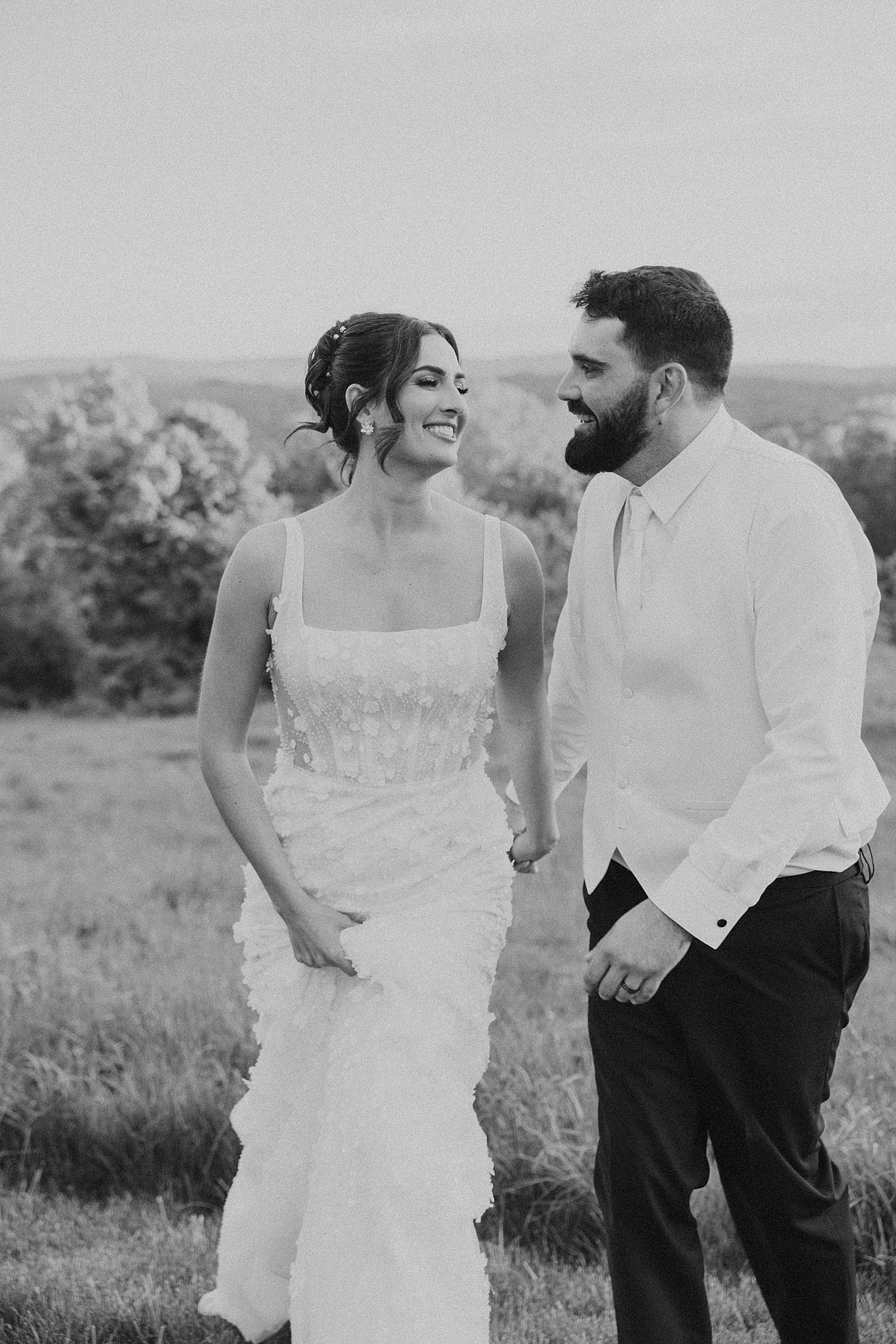 Black and white photo of a smiling bride in a ruffled gown holding hands with her groom.