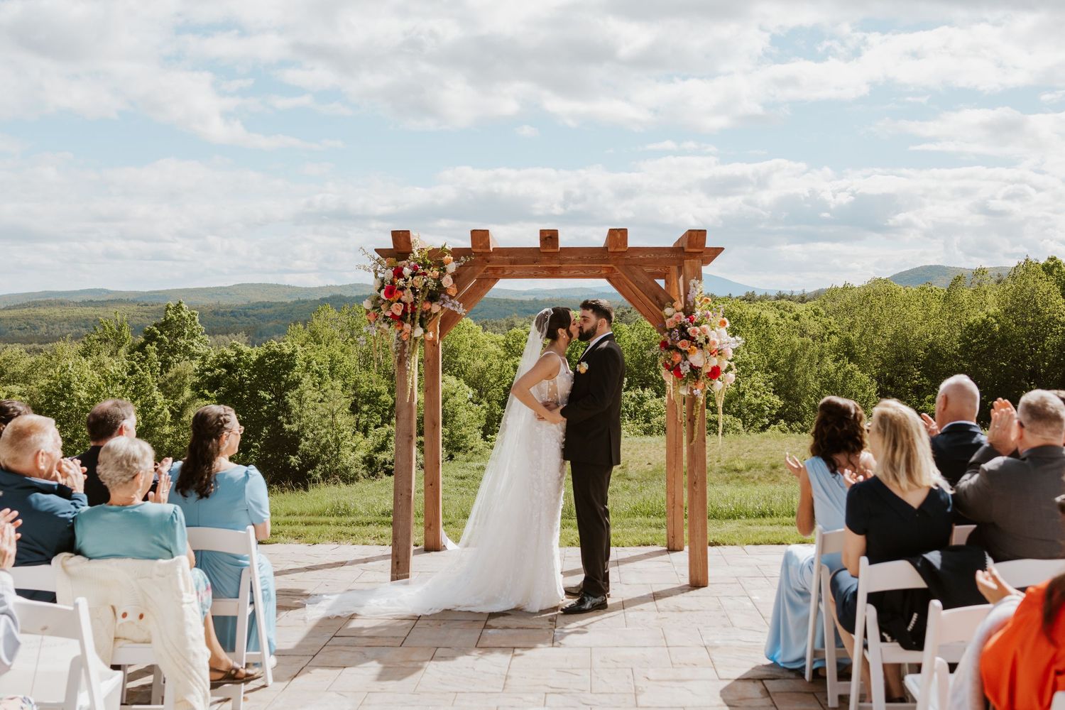 Couple kissing at outdoor wedding ceremony under wooden arch with floral arrangements and scenic mountain views.