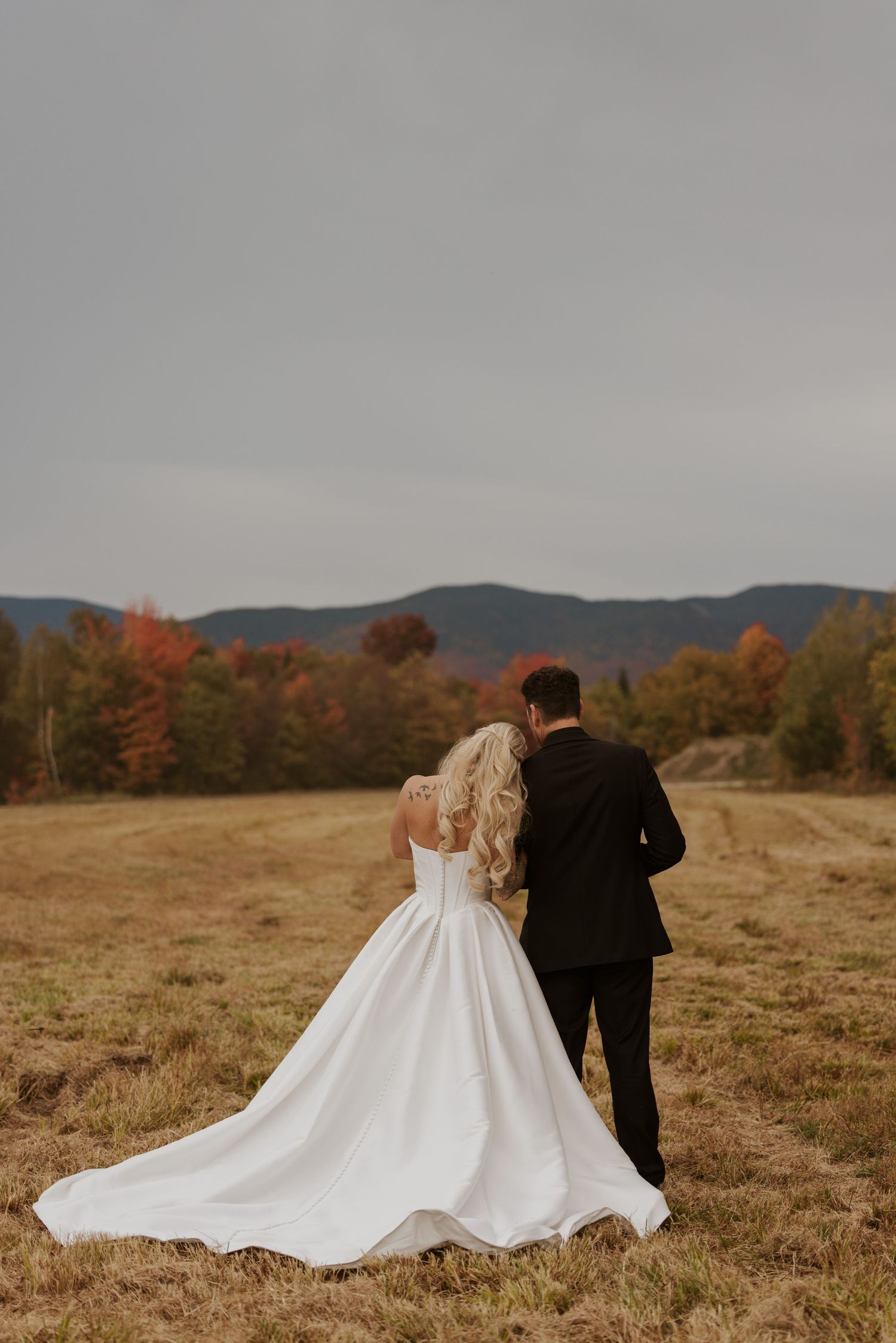 Bride and groom walking in autumn field with colorful foliage and mountains in background on wedding day.