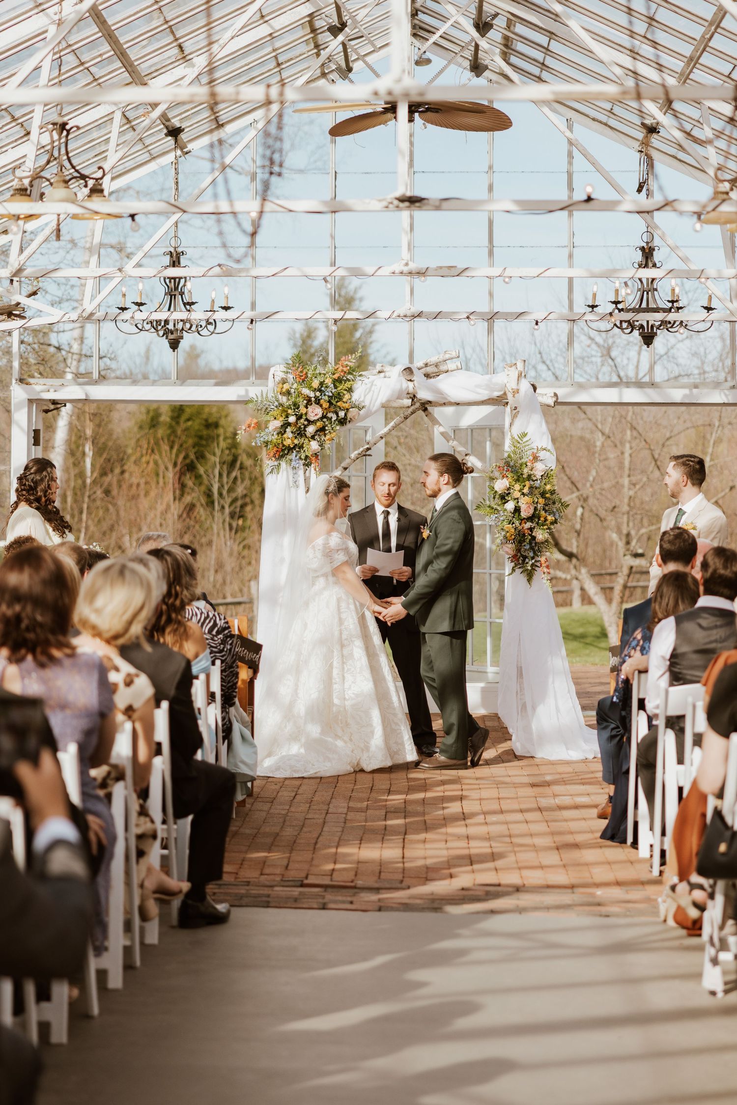 Wedding ceremony in glass greenhouse venue with brick aisle, floral arch, and seated guests.