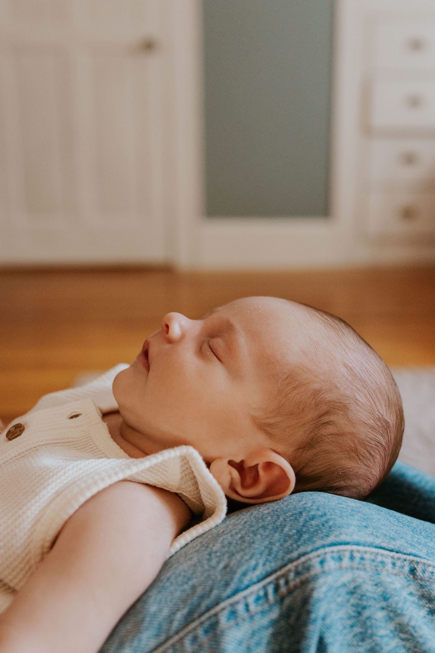 Serene sleeping newborn baby in cream ribbed onesie resting on parent's lap in softly lit nursery.