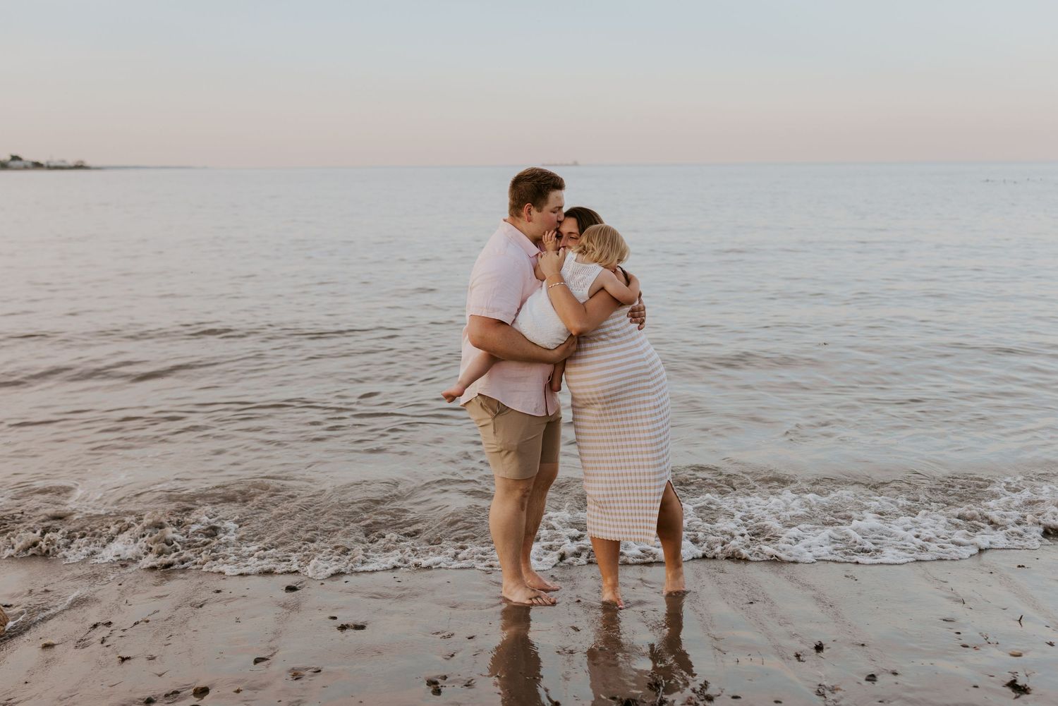 Couple embracing on beach at sunset during maternity photoshoot with ocean waves behind them.
