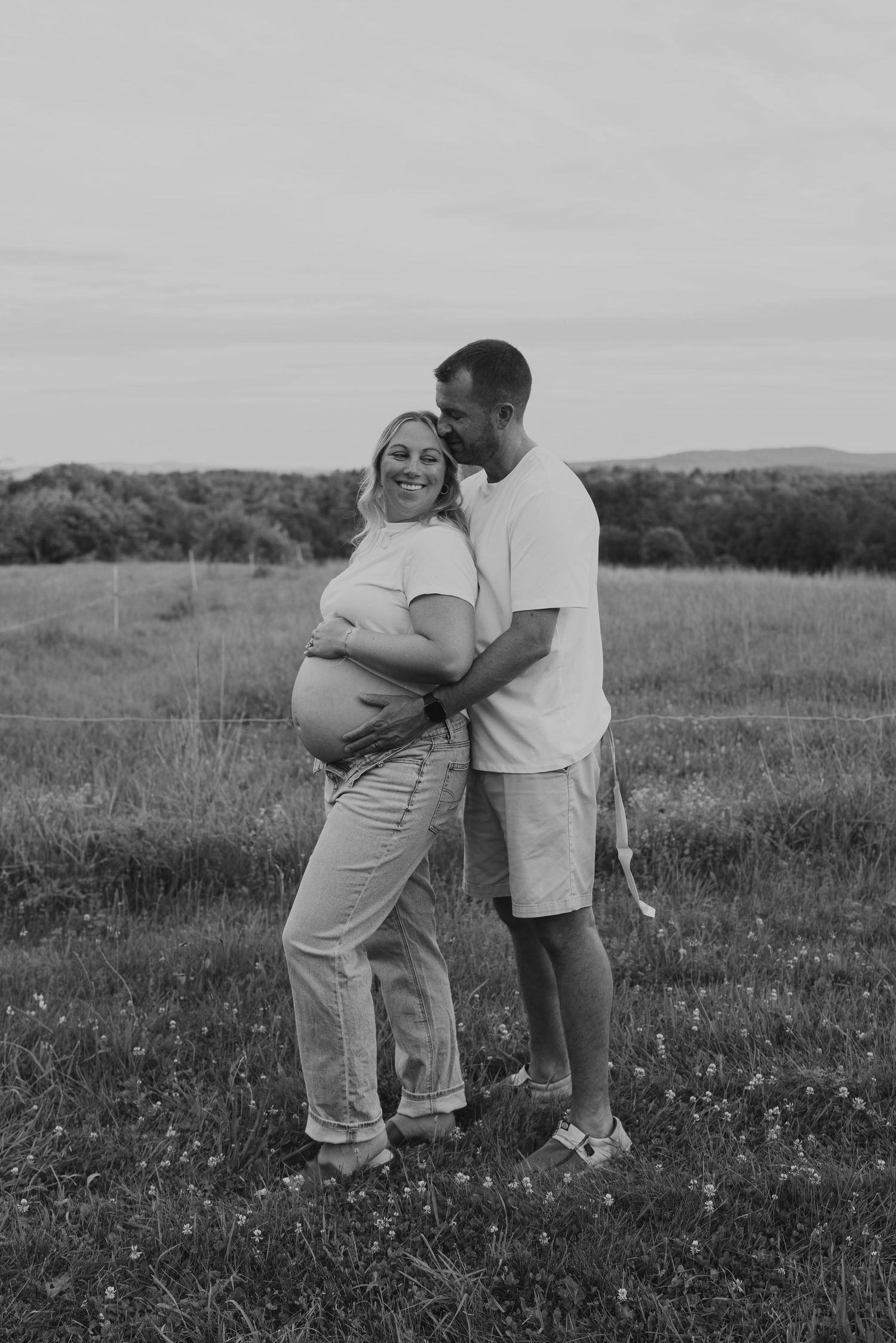 Black and white maternity photo of a couple embracing in a field, man kissing pregnant woman's head tenderly.
