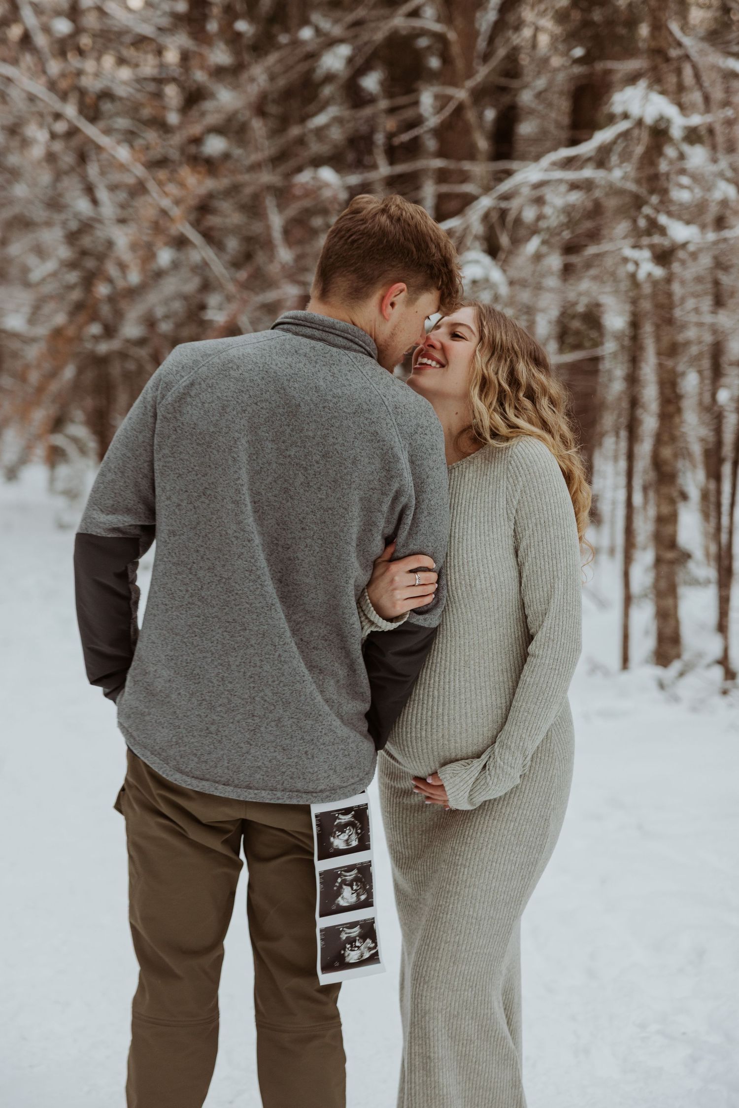 Joyful expecting couple laughing together in snowy woods, holding ultrasound photos between them.
