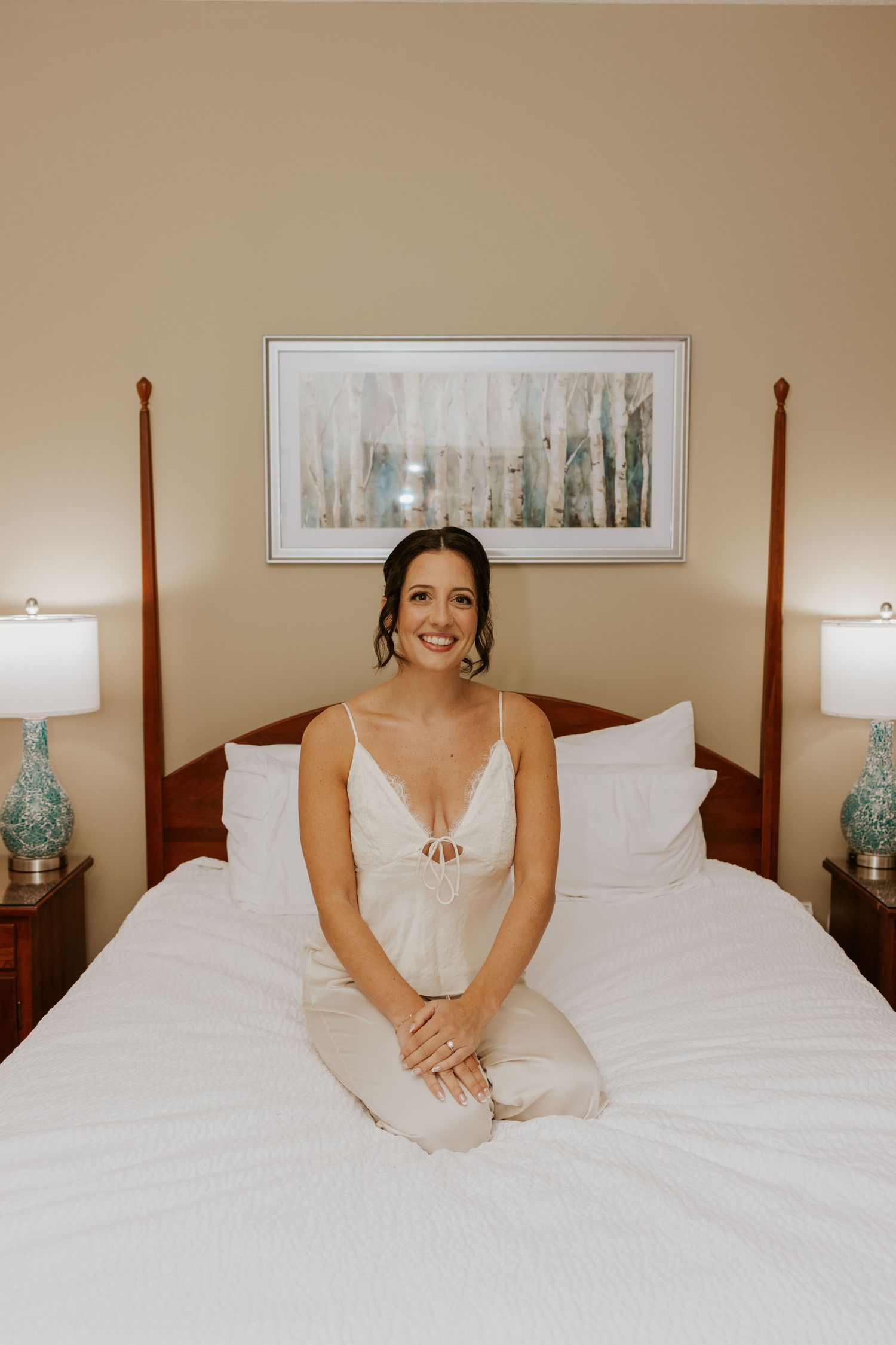 Smiling bride in white slip dress sits on a hotel bed before her wedding ceremony.