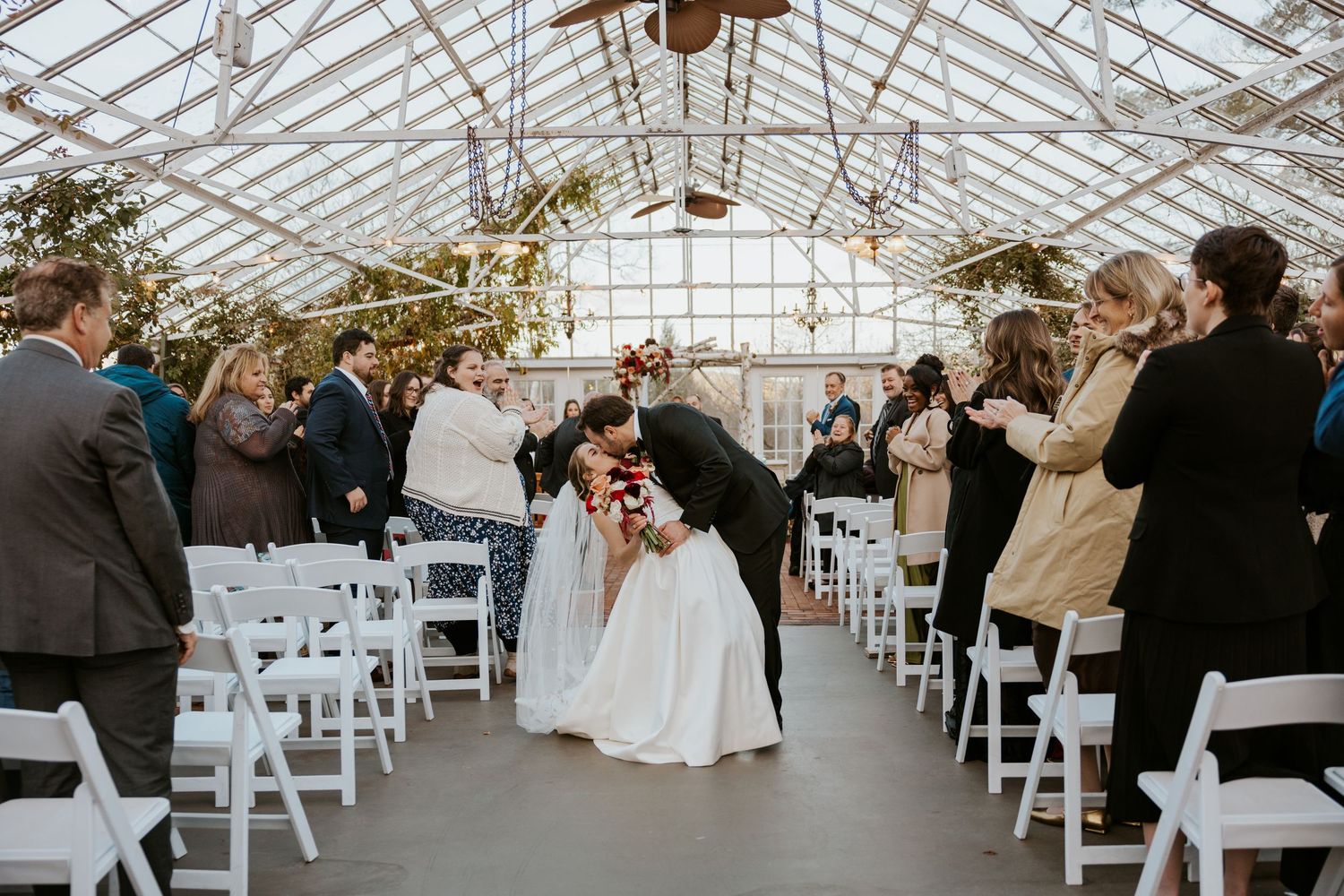 Bride and groom kiss in the aisle as wedding guests celebrate in a stunning glass greenhouse venue.