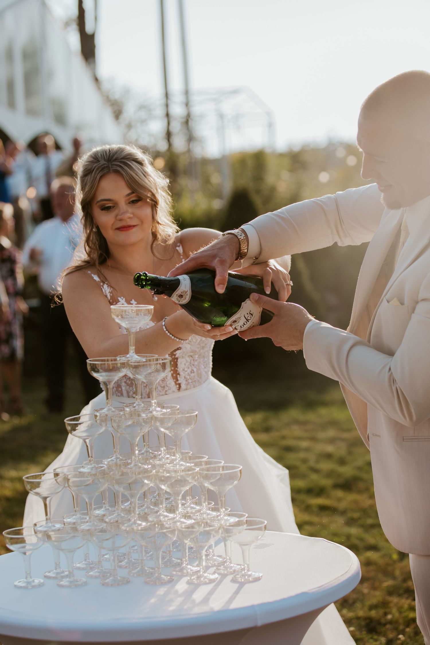 Bride and groom pouring champagne tower at outdoor wedding reception in golden sunlight.