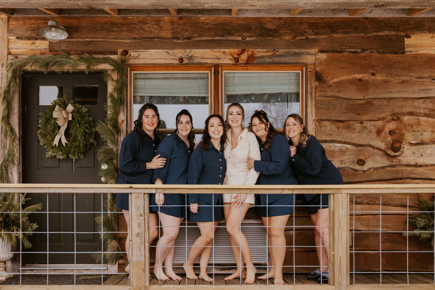 Bride and bridesmaids in navy robes smiling together on a rustic log cabin porch decorated with greenery.