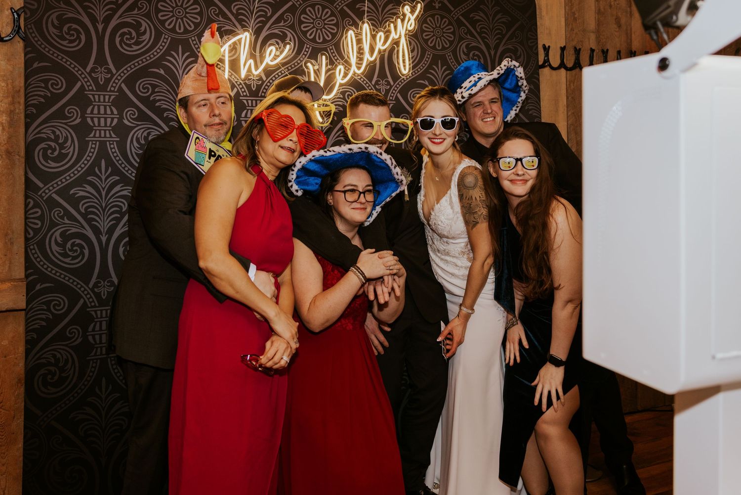 Group of friends posing with fun props at a wedding photo booth in front of a decorative wall.
