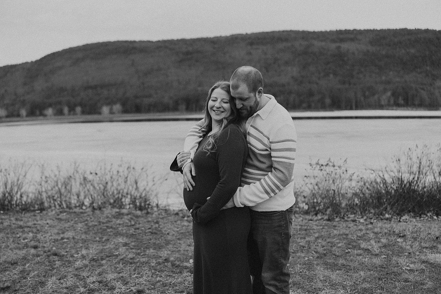 Black and white photo of a couple embracing outdoors near a lake with rolling hills in the background.