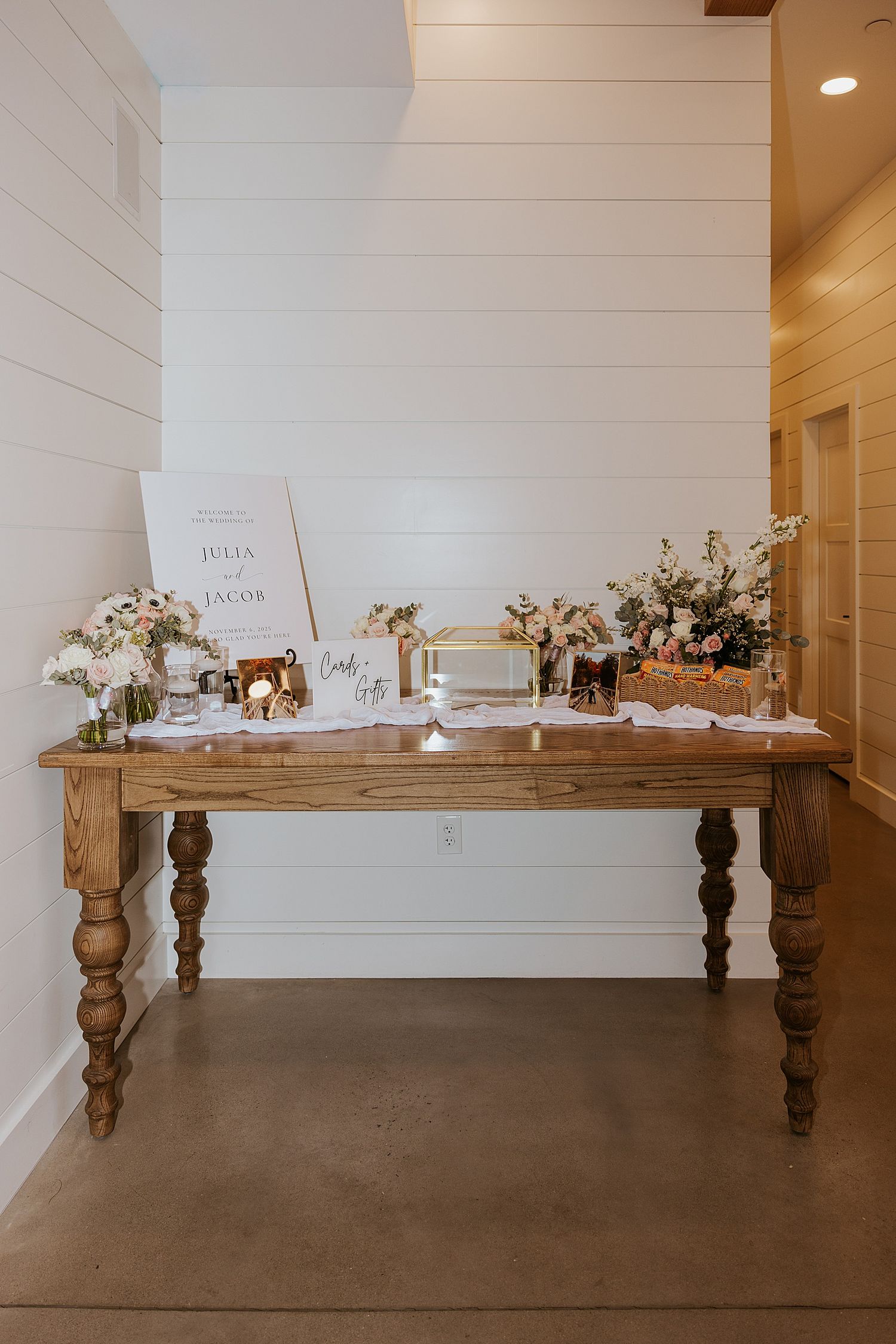 Rustic wooden table with wedding welcome sign, floral arrangements, and gift display in a white shiplap venue.