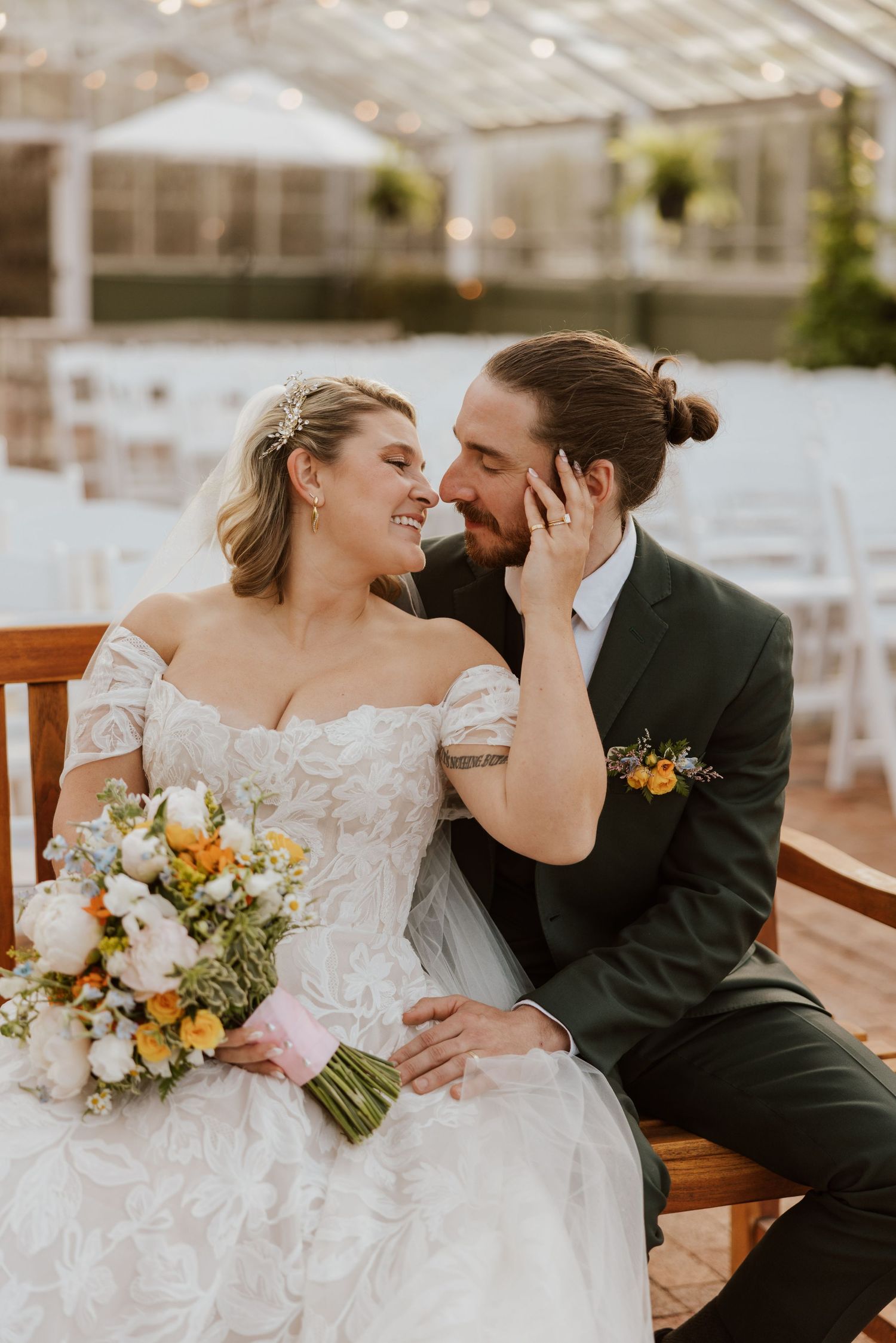 Smiling bride and groom share intimate moment seated near greenhouse wedding venue.