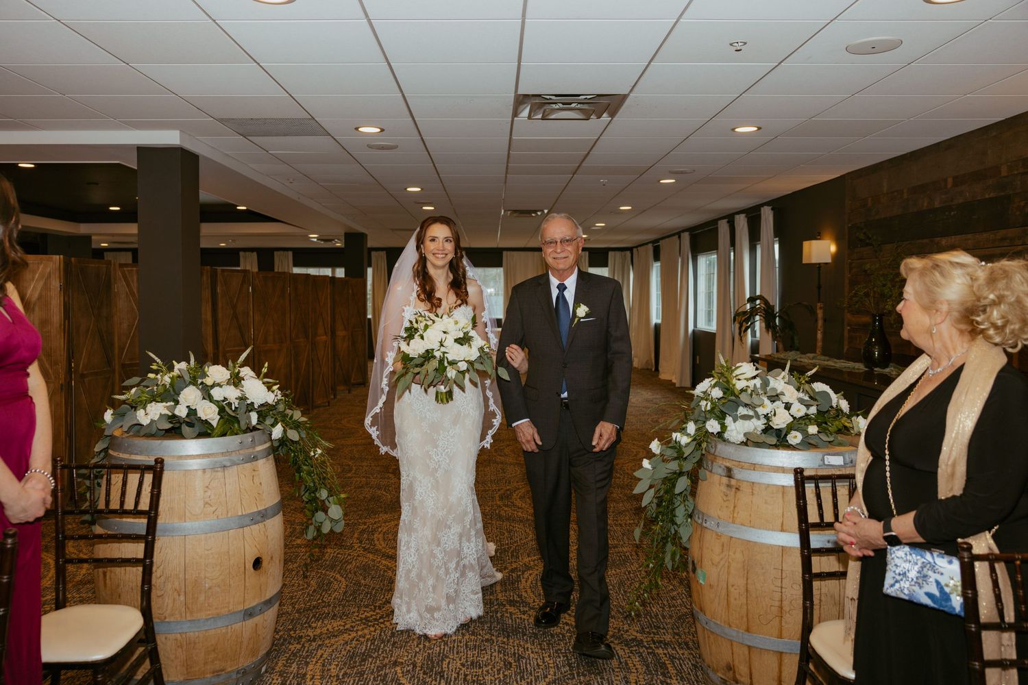 A wedding ceremony setup with rustic wine barrels and white floral arrangements lining the aisle in an indoor venue.